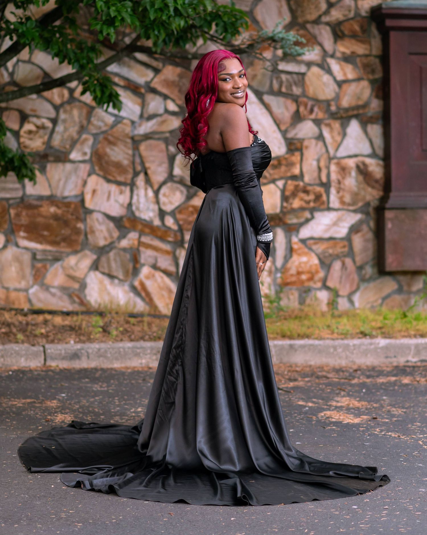 Person in flowing black gown with red hair poses elegantly against stone wall backdrop.