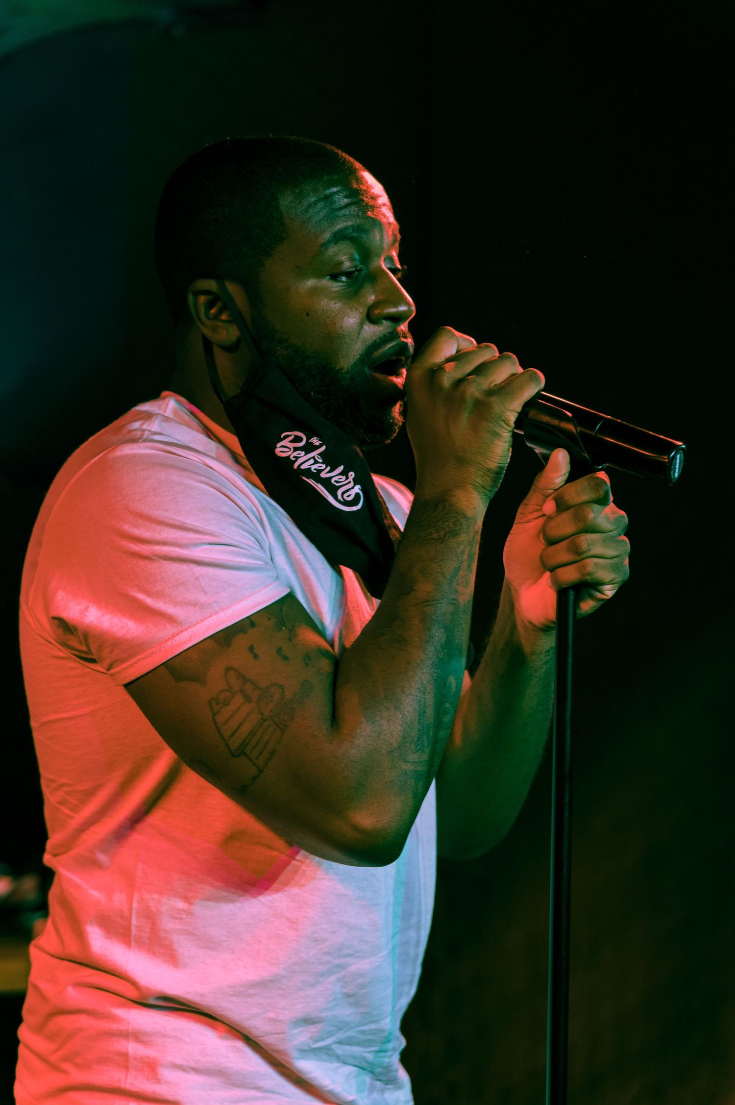 A performer in a pink t-shirt sings passionately into a microphone during a dimly lit concert.