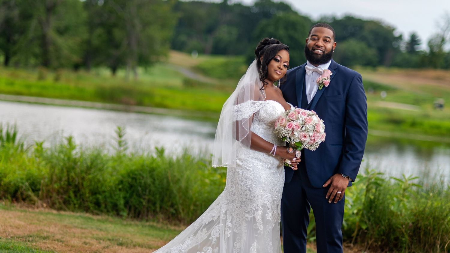 Newlyweds pose by lakeside with bride holding bouquet in elegant wedding attire.