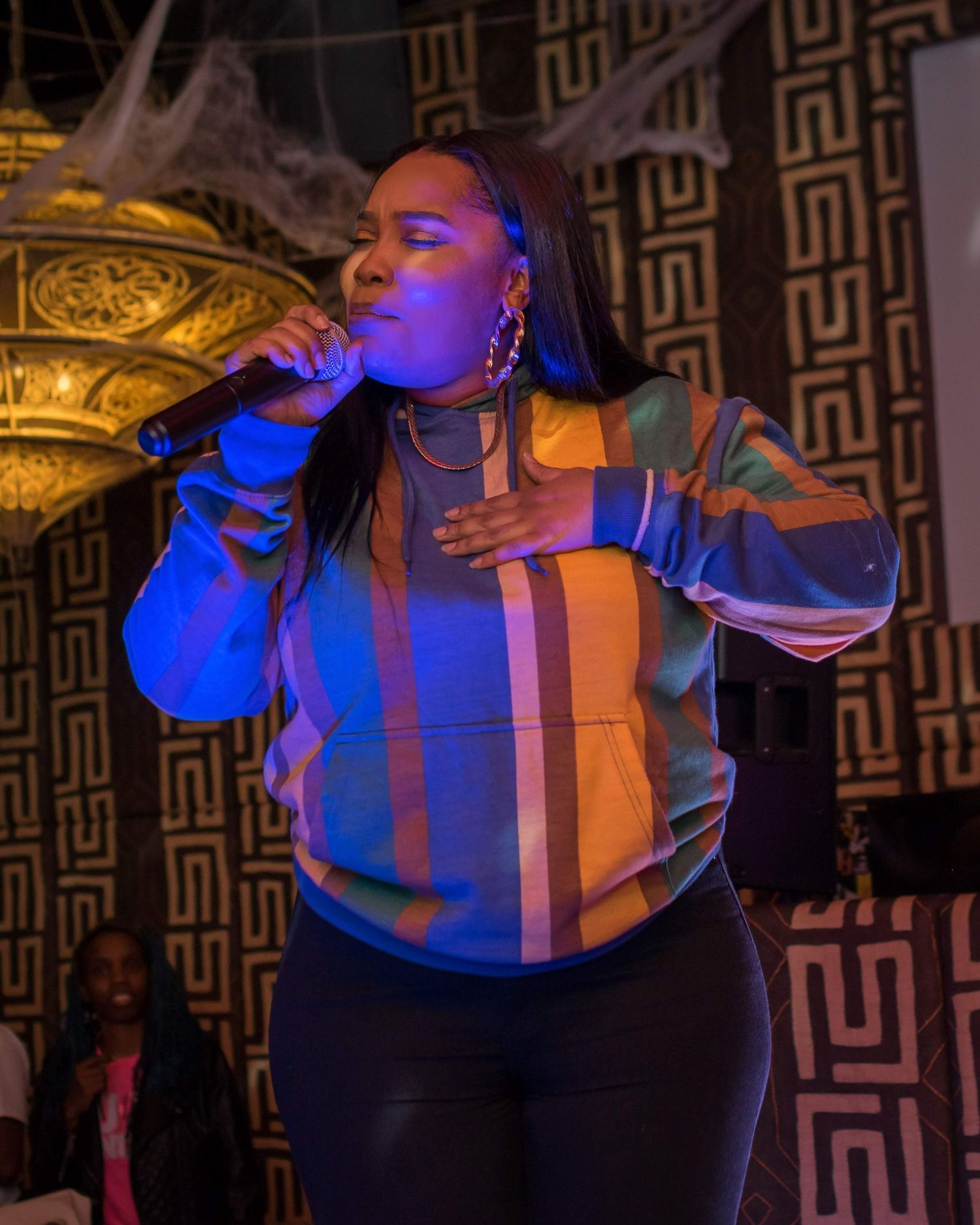 A performer in a striped sweater sings emotionally under blue stage lighting against a patterned backdrop.