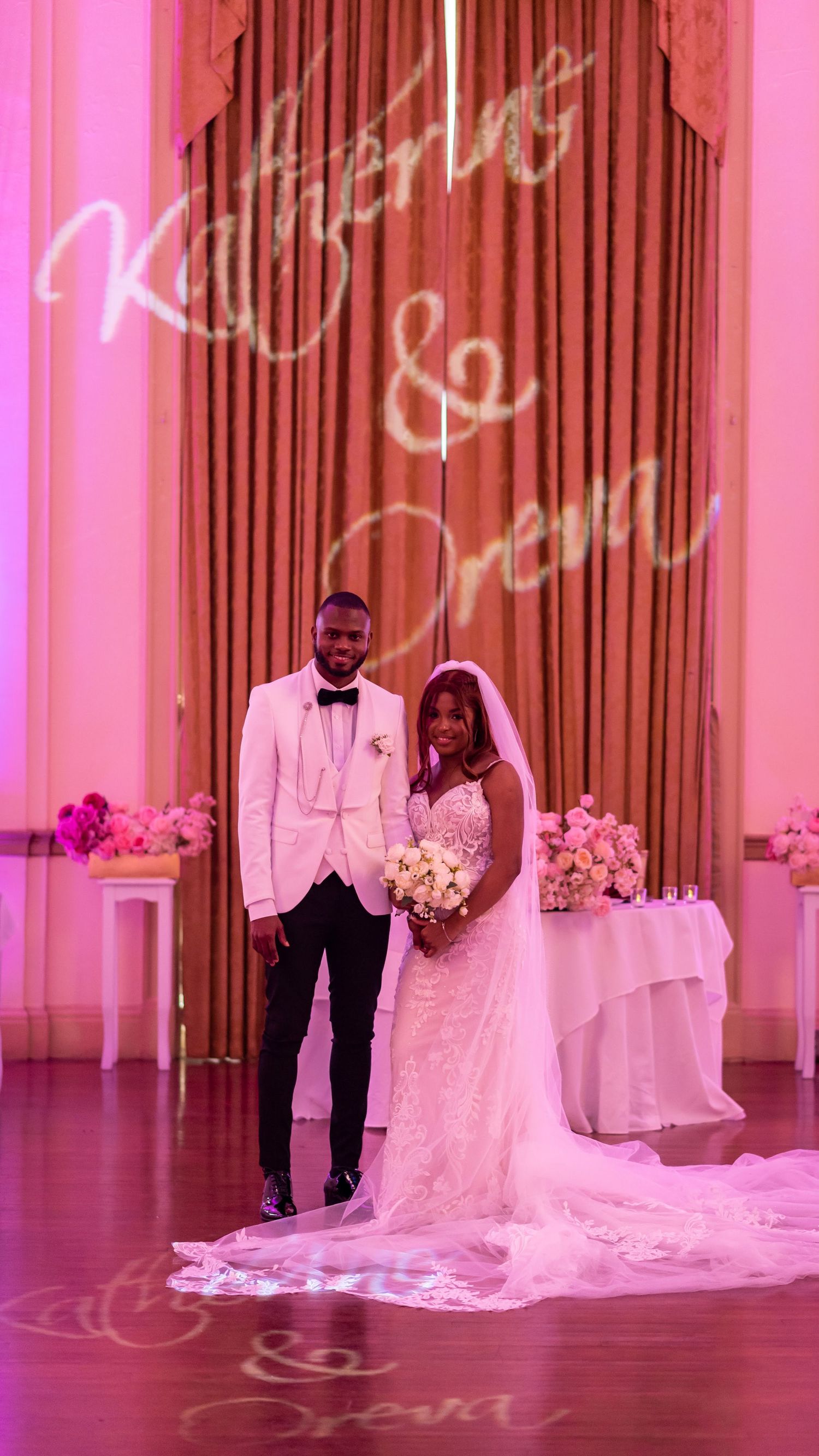 A couple stands beneath elegant decor at their wedding reception with pink lighting and ornate wall details.
