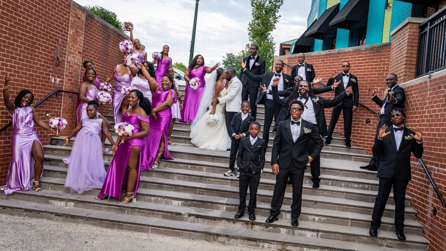 Wedding party poses on brick stairs with bridesmaids in purple dresses and groomsmen in black suits.