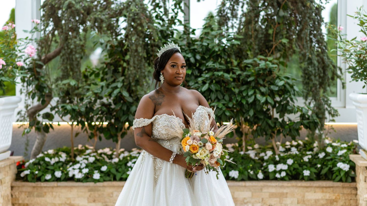 A bride in an off-shoulder wedding gown holding a vibrant bouquet against a floral garden backdrop.