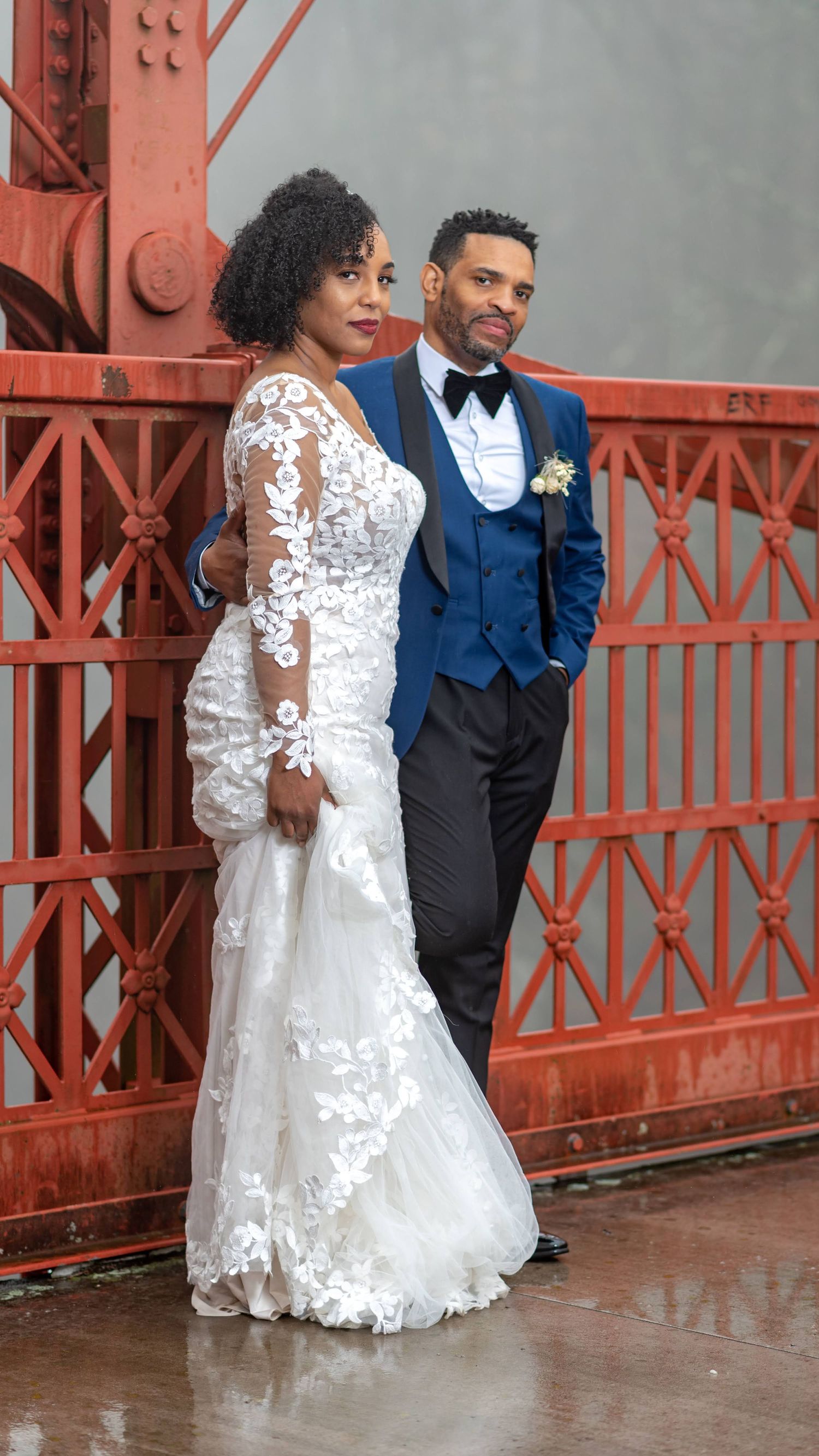 Wedding couple poses against red industrial bridge structure in formal attire.