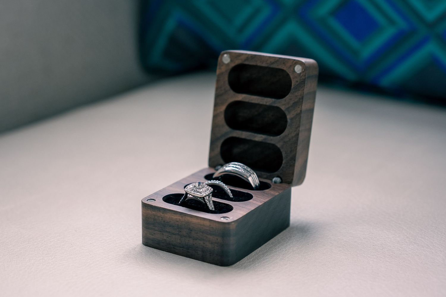 Elegant wooden ring box with three slots holding diamond engagement ring and wedding bands on white surface.