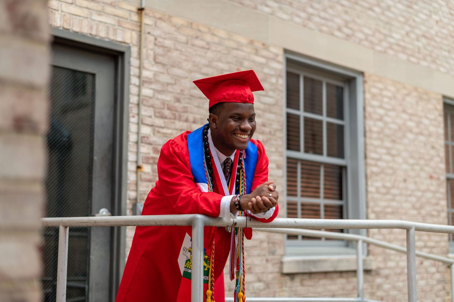 A graduate in red cap and gown smiles while leaning on a railing outside a brick building.