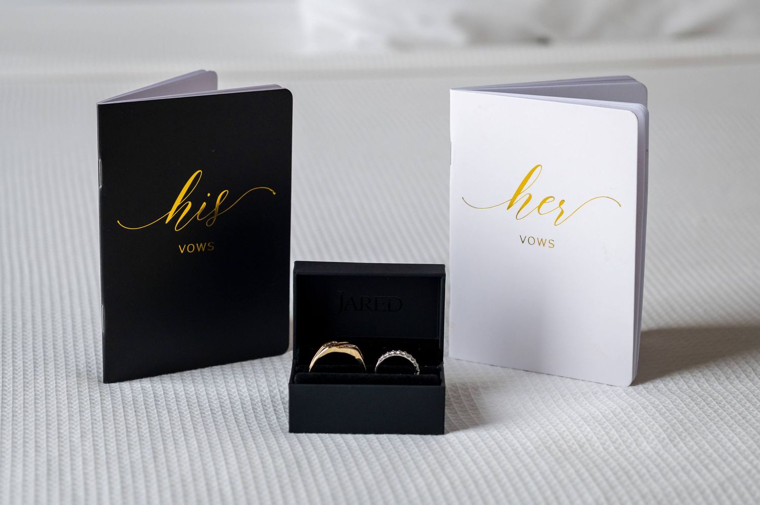 Black and white vow books with gold lettering beside wedding rings in a box.