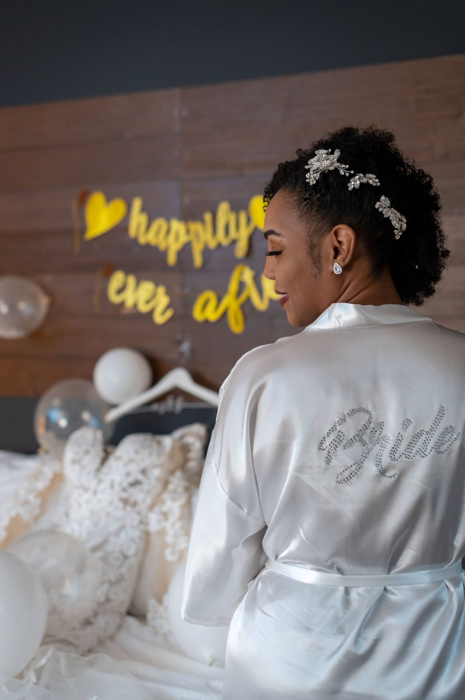 A bride in a white satin robe with rhinestone details poses in front of a 'happily ever after' sign.