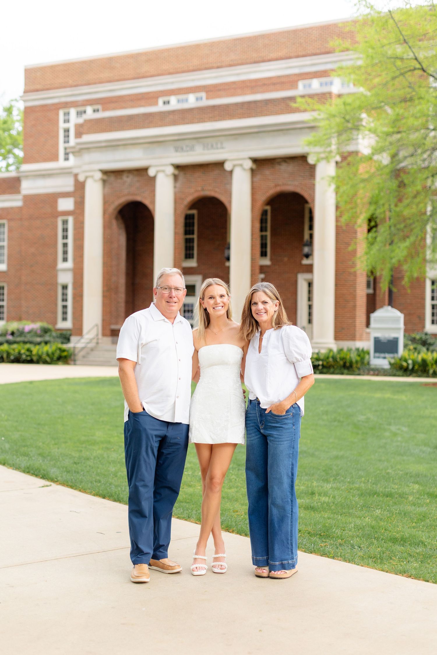 Three people pose together on a grassy lawn in front of a brick colonial-style building with white columns.