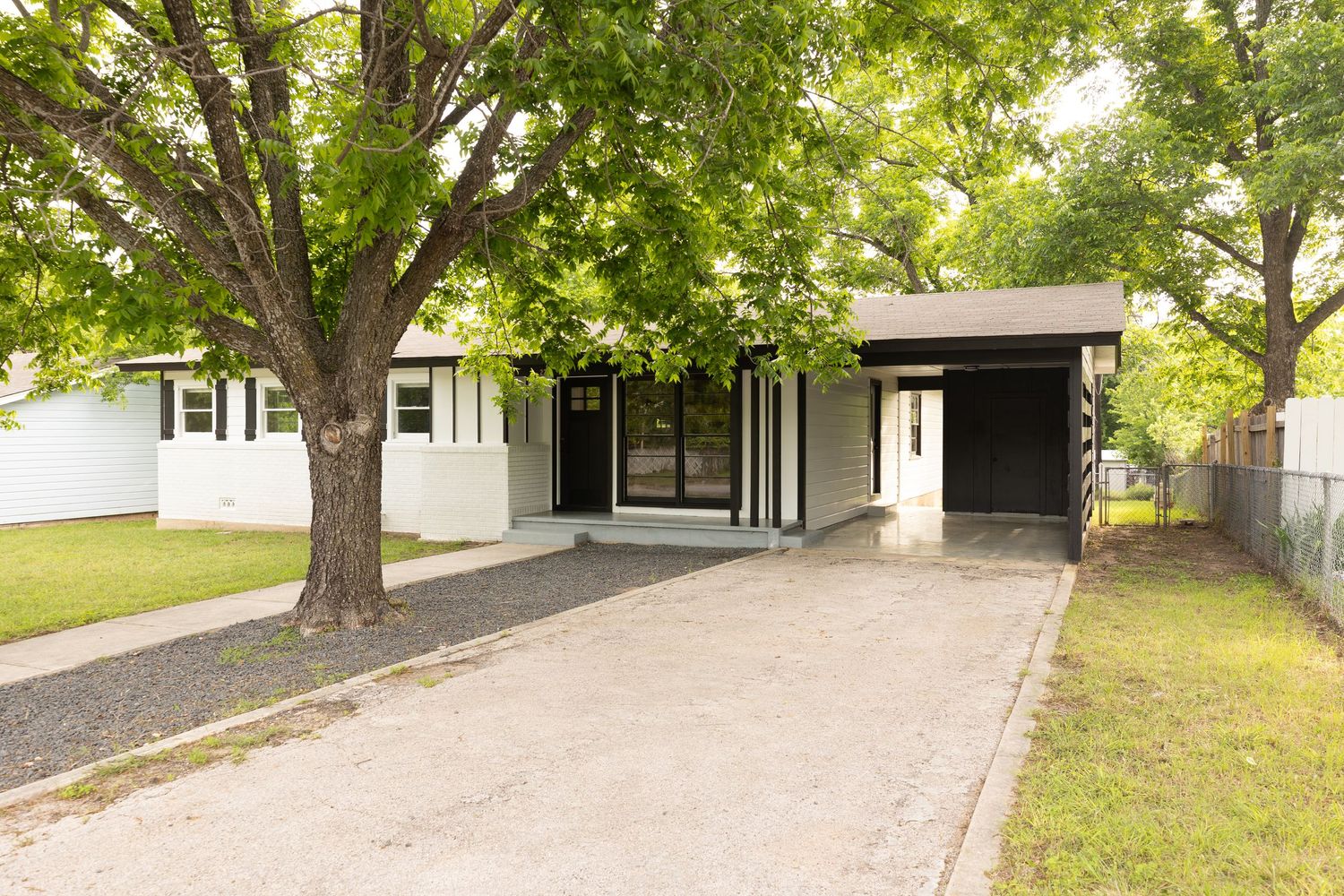 Single-story ranch home with mature shade trees, white exterior, covered carport, and concrete driveway on a green lawn.