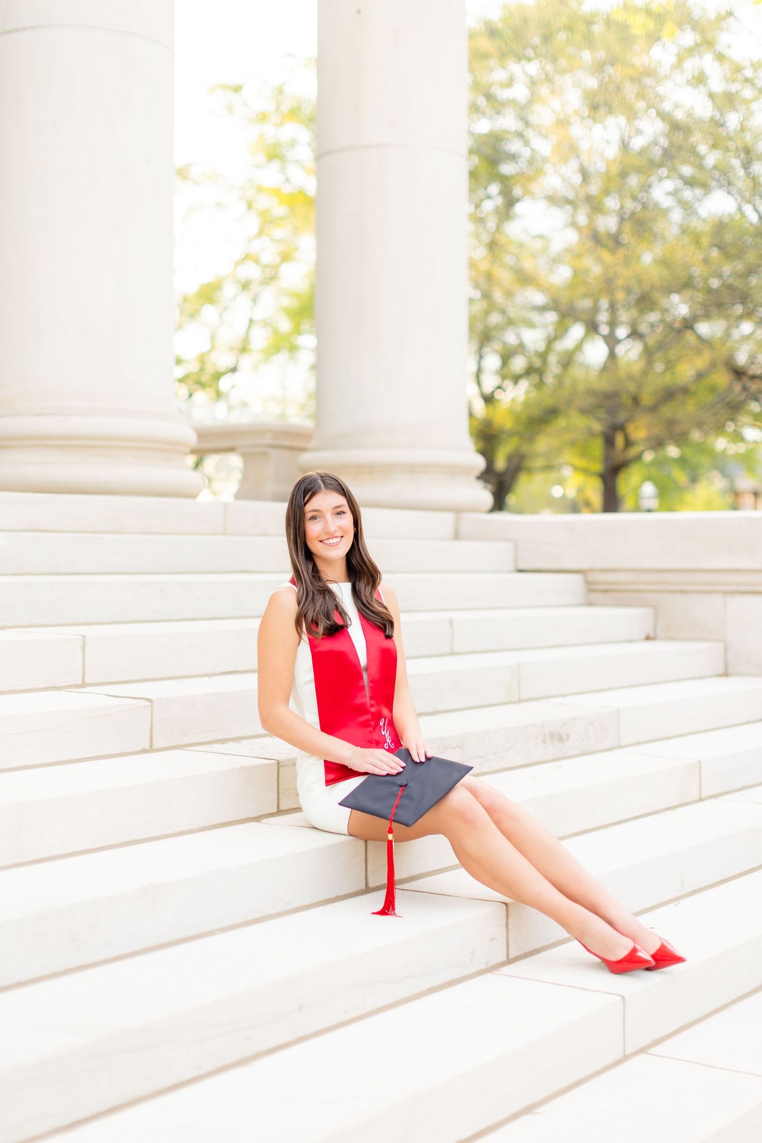 A person in a red dress and matching shoes sits gracefully on white marble steps in an outdoor setting.
