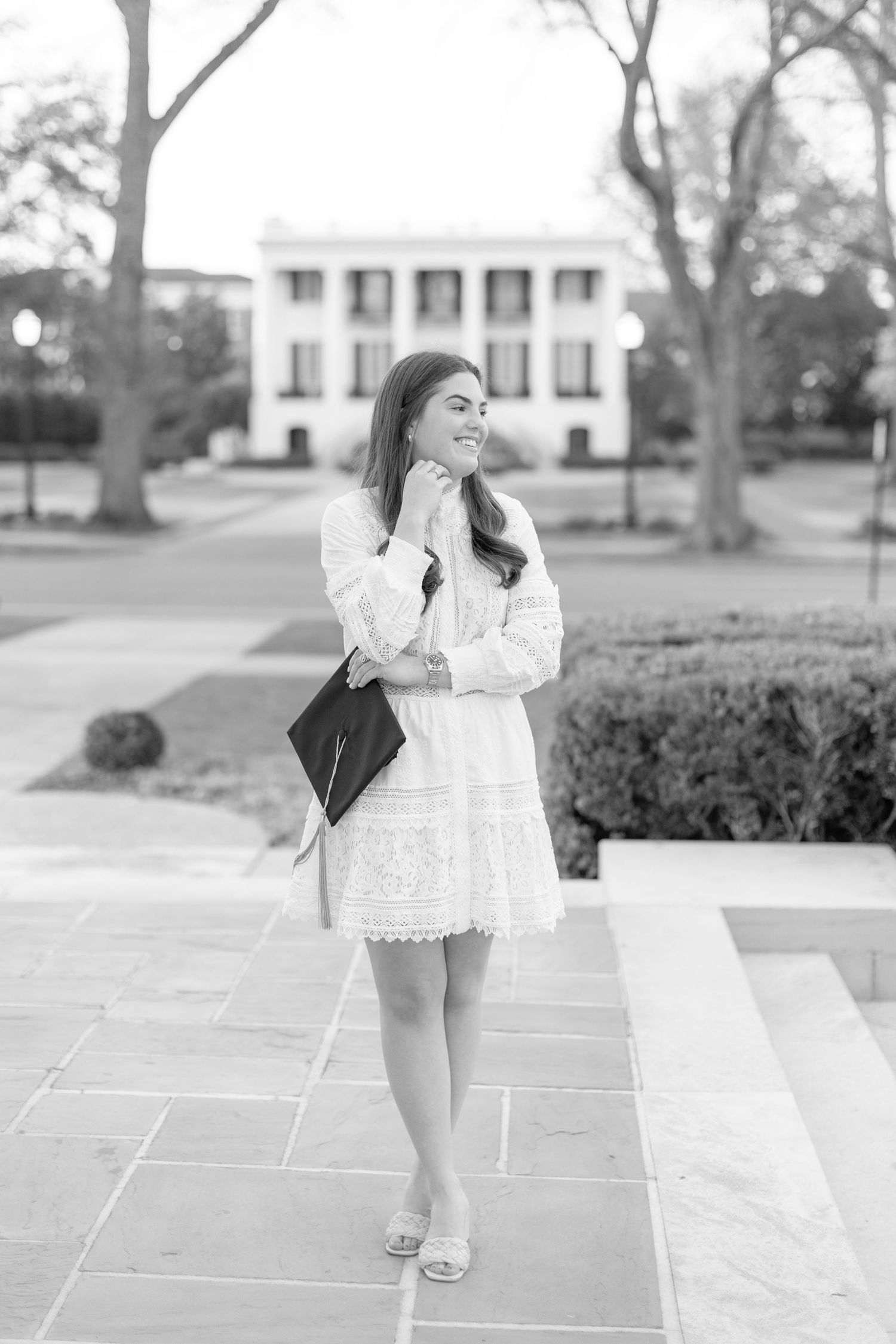 Black and white photo of someone in a white lace dress posing outdoors in front of a stately white building.
