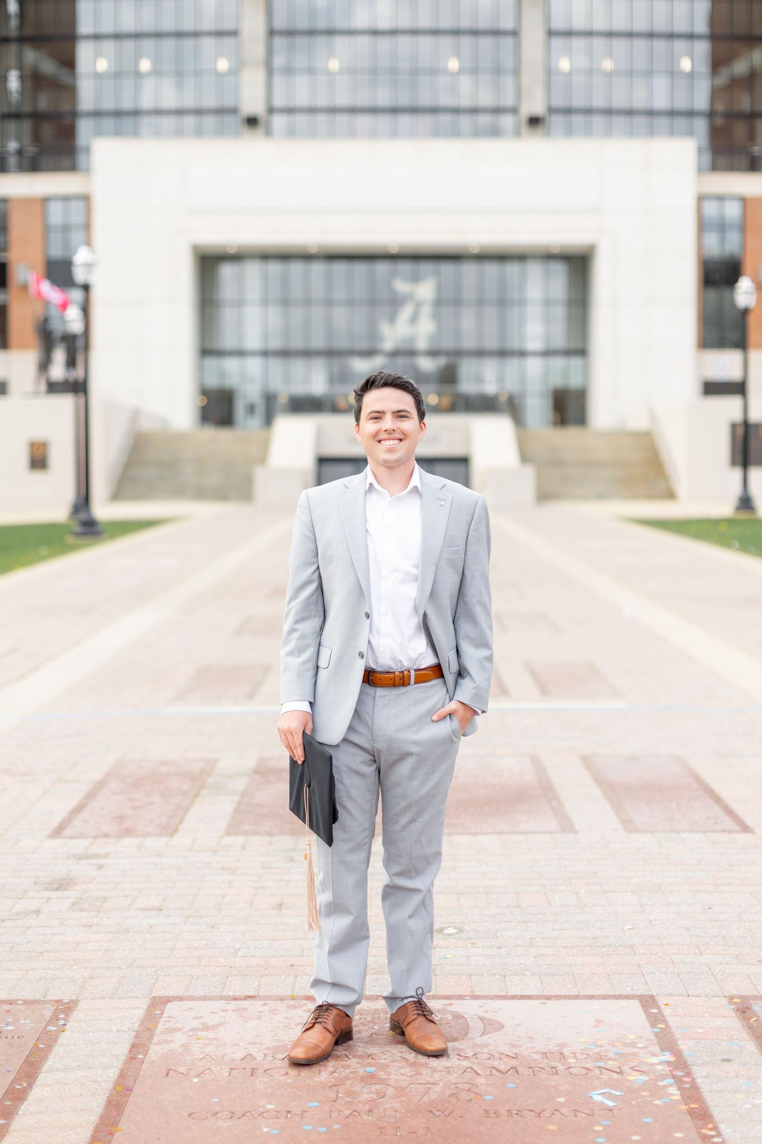 A person in a light gray suit and brown shoes stands on a patterned walkway in front of a modern building entrance.