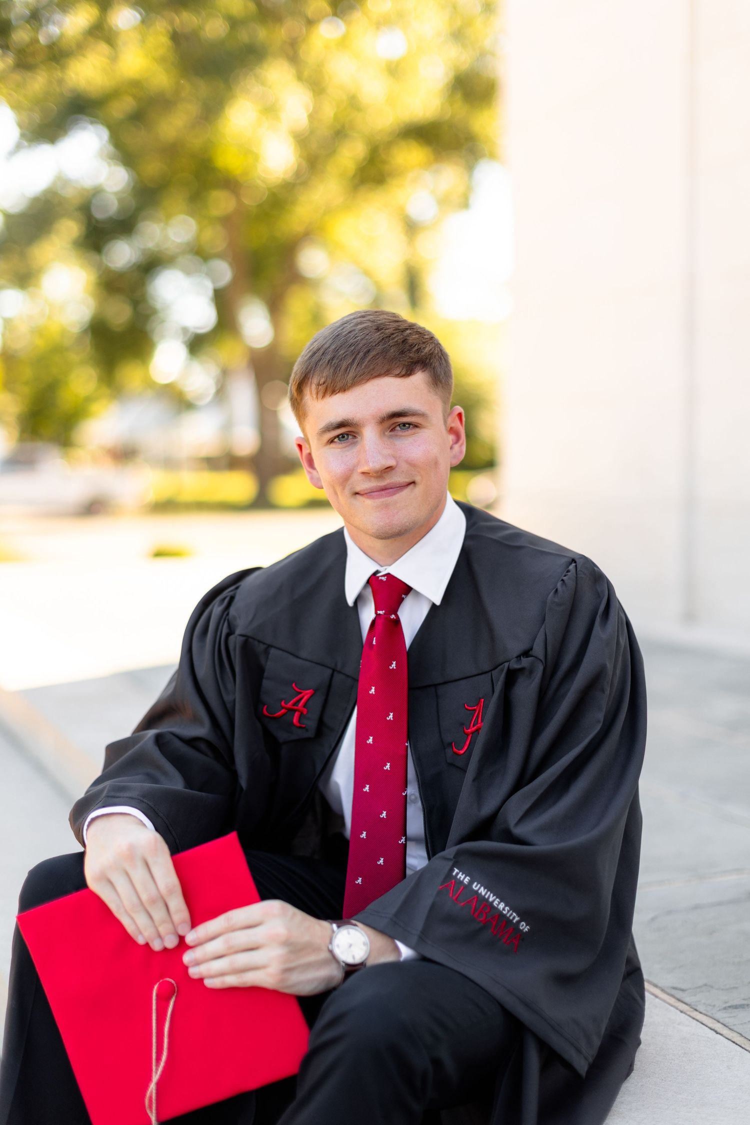 Graduate in black cap and gown holds red diploma folder while posing against white columns in sunlight.