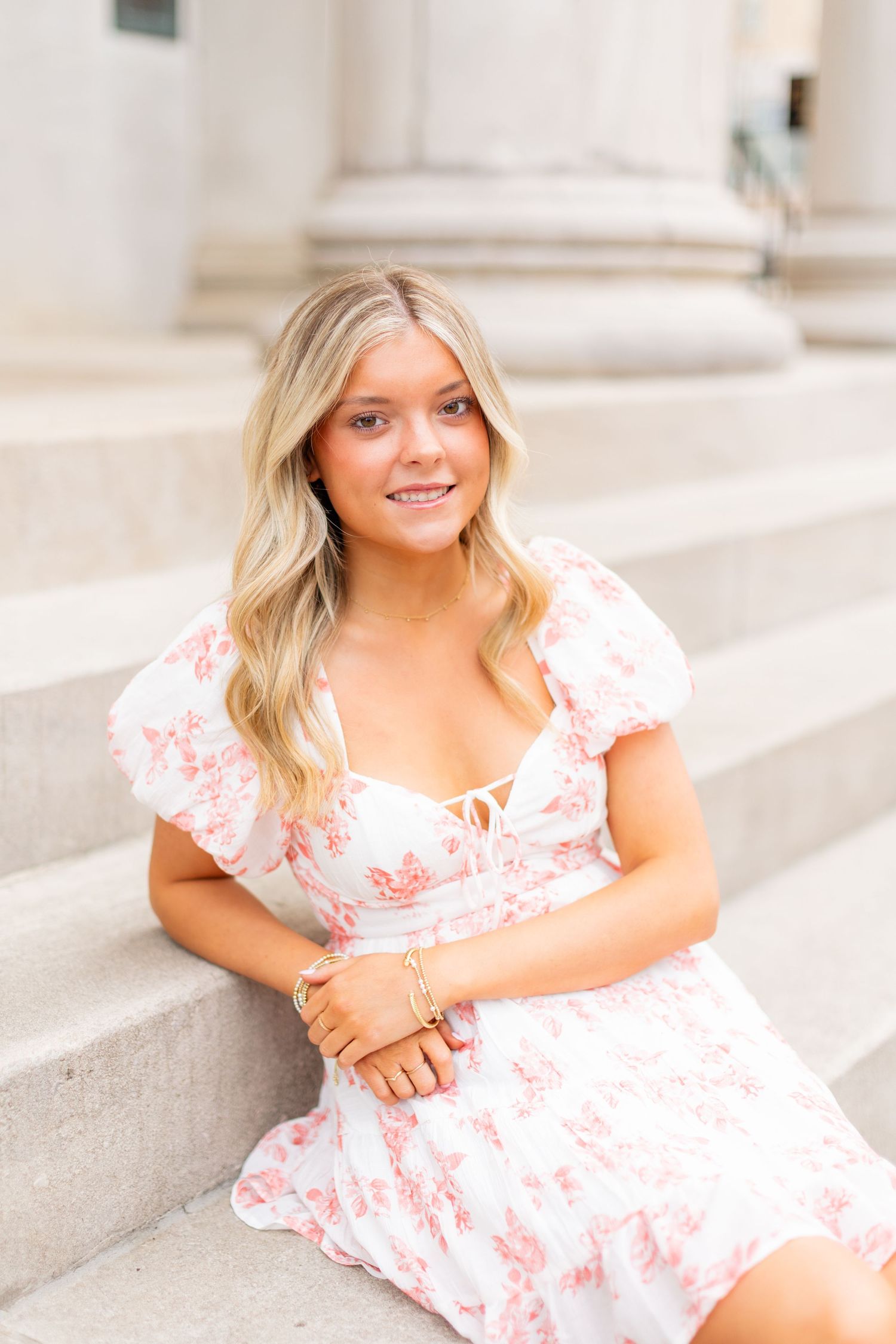 A person in a white and pink floral dress sits on stone steps in a bright outdoor setting.