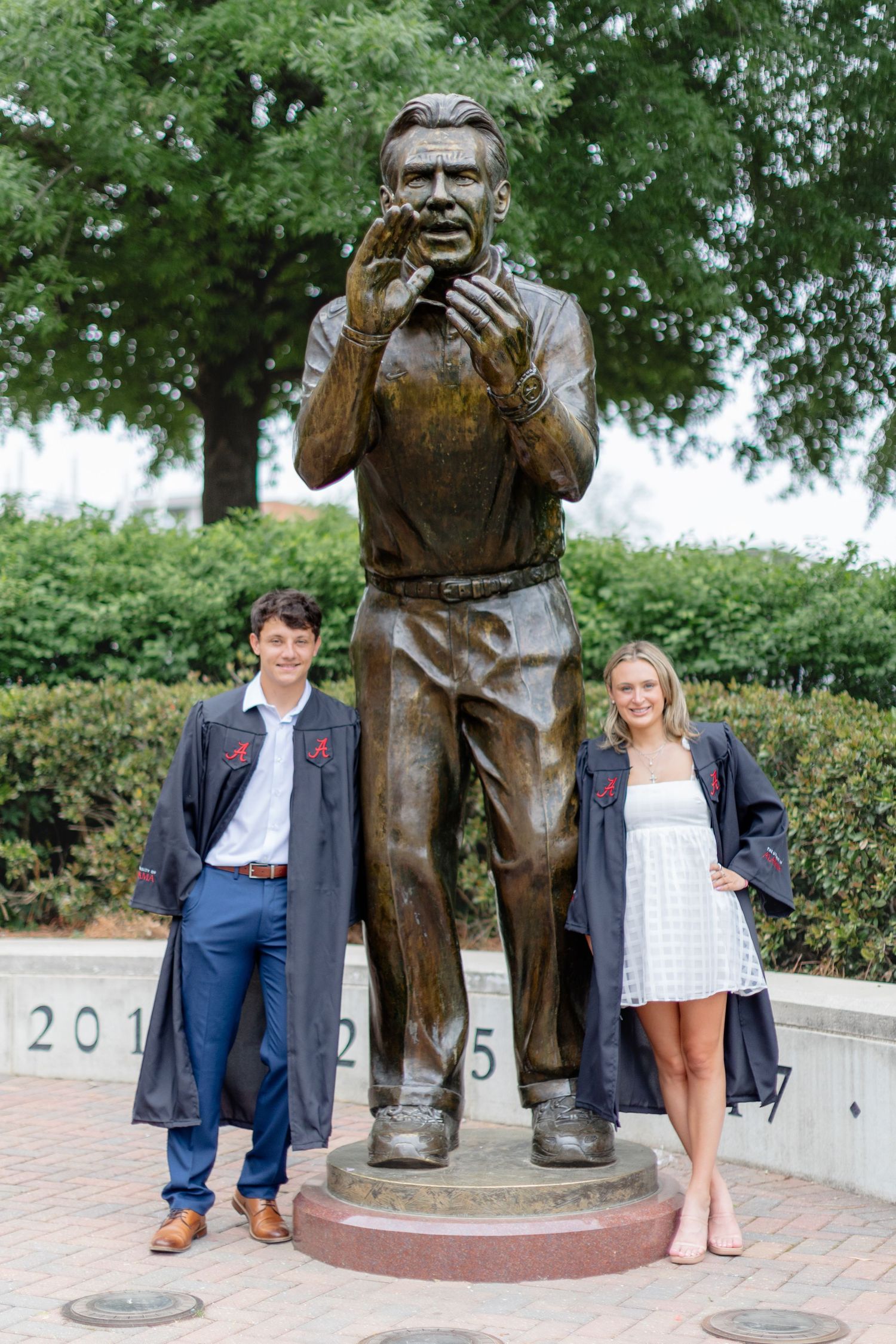 Two graduates pose with statue outside brick building wearing graduation gowns.