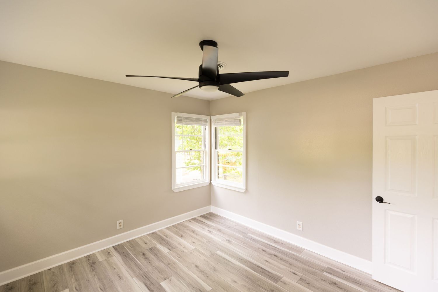 Bright empty bedroom featuring a ceiling fan, corner windows, light hardwood floors, and neutral gray walls.
