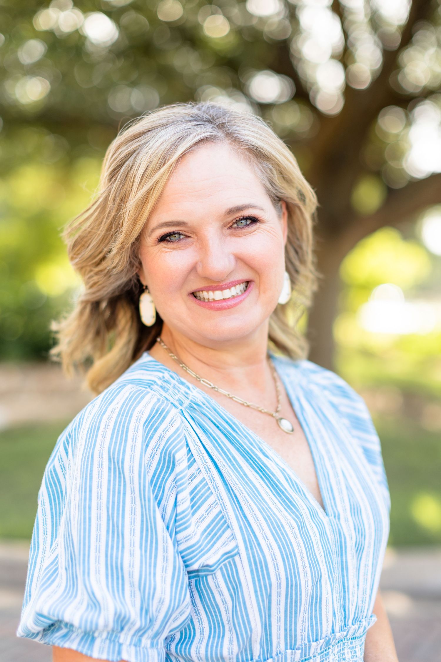 A person wearing a blue and white striped blouse smiles warmly in an outdoor setting with soft bokeh lighting effects.