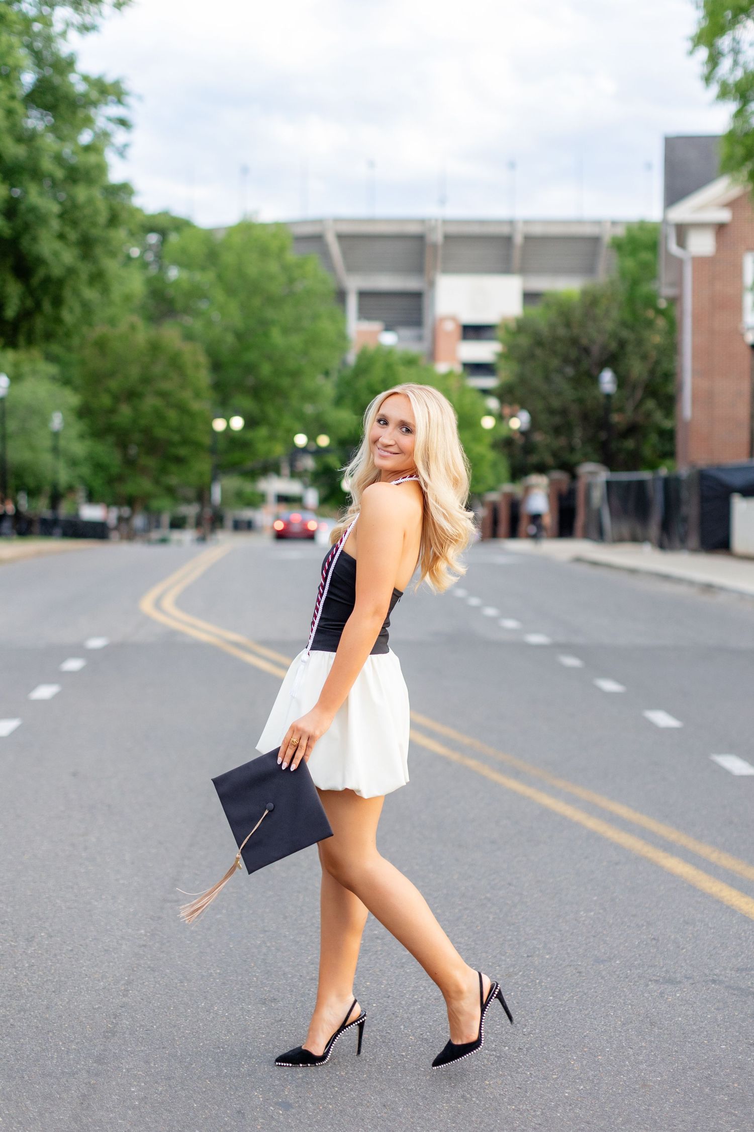 Fashionable figure in white skirt and black top walking confidently down an urban street with graduation cap.