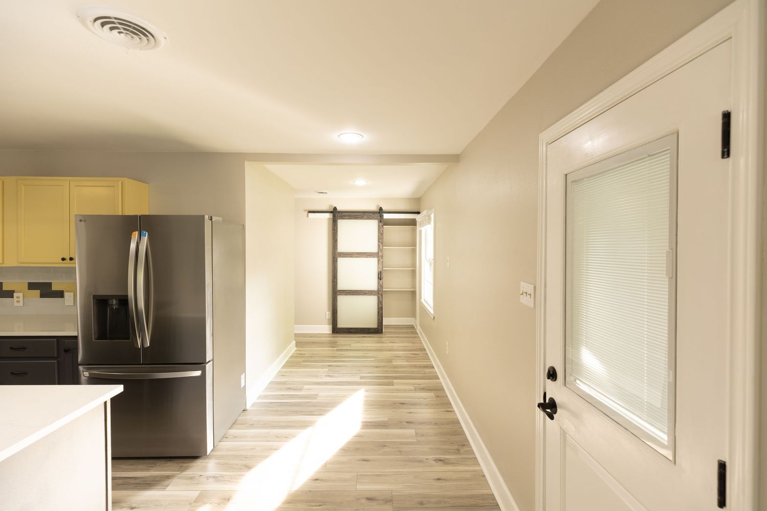 Modern kitchen hallway with stainless steel refrigerator, yellow cabinets, barn door, and light hardwood floors.
