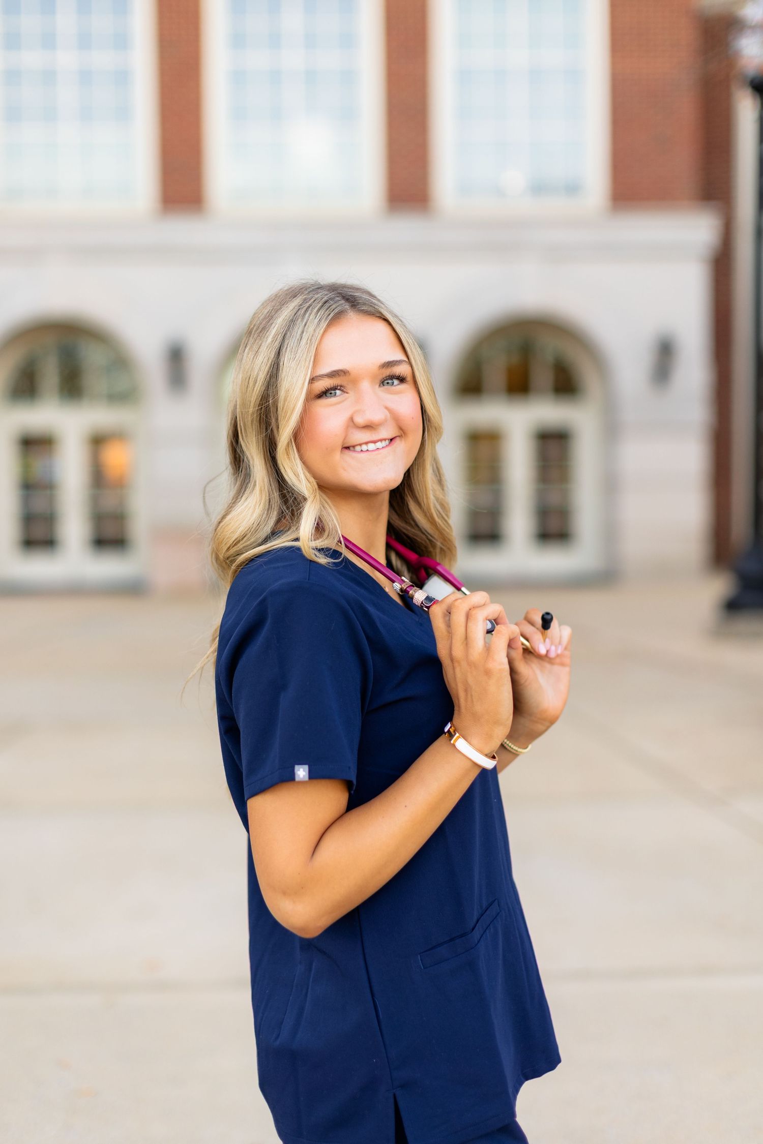 Healthcare professional in navy blue scrubs smiles while standing outside a building with arched windows.