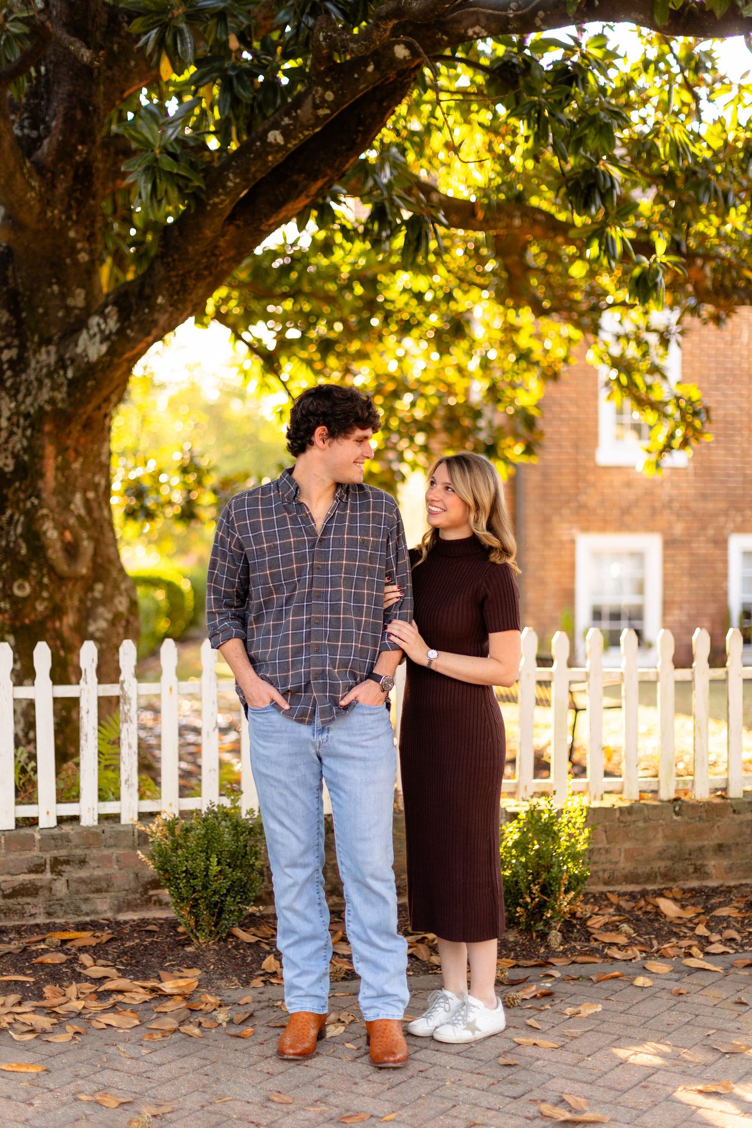 A couple shares romantic moments in front of a white picket fence and brick wall on a sunny autumn day.