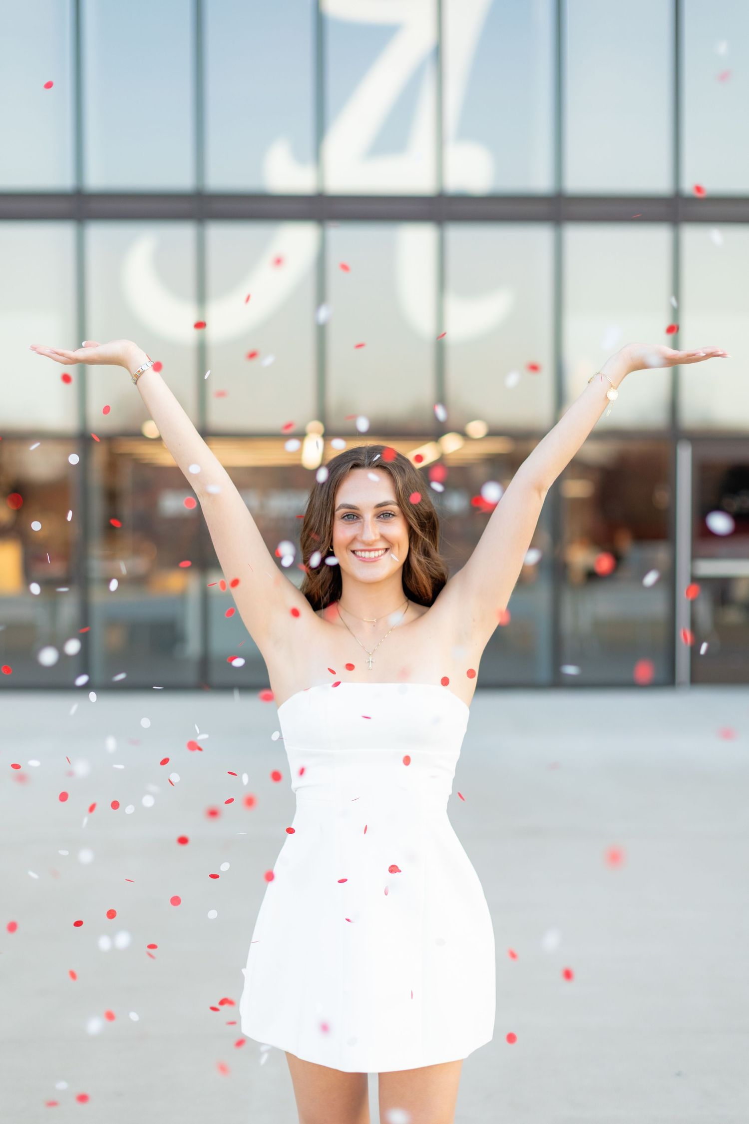 Graduate in white dress celebrates with arms raised as red confetti falls.