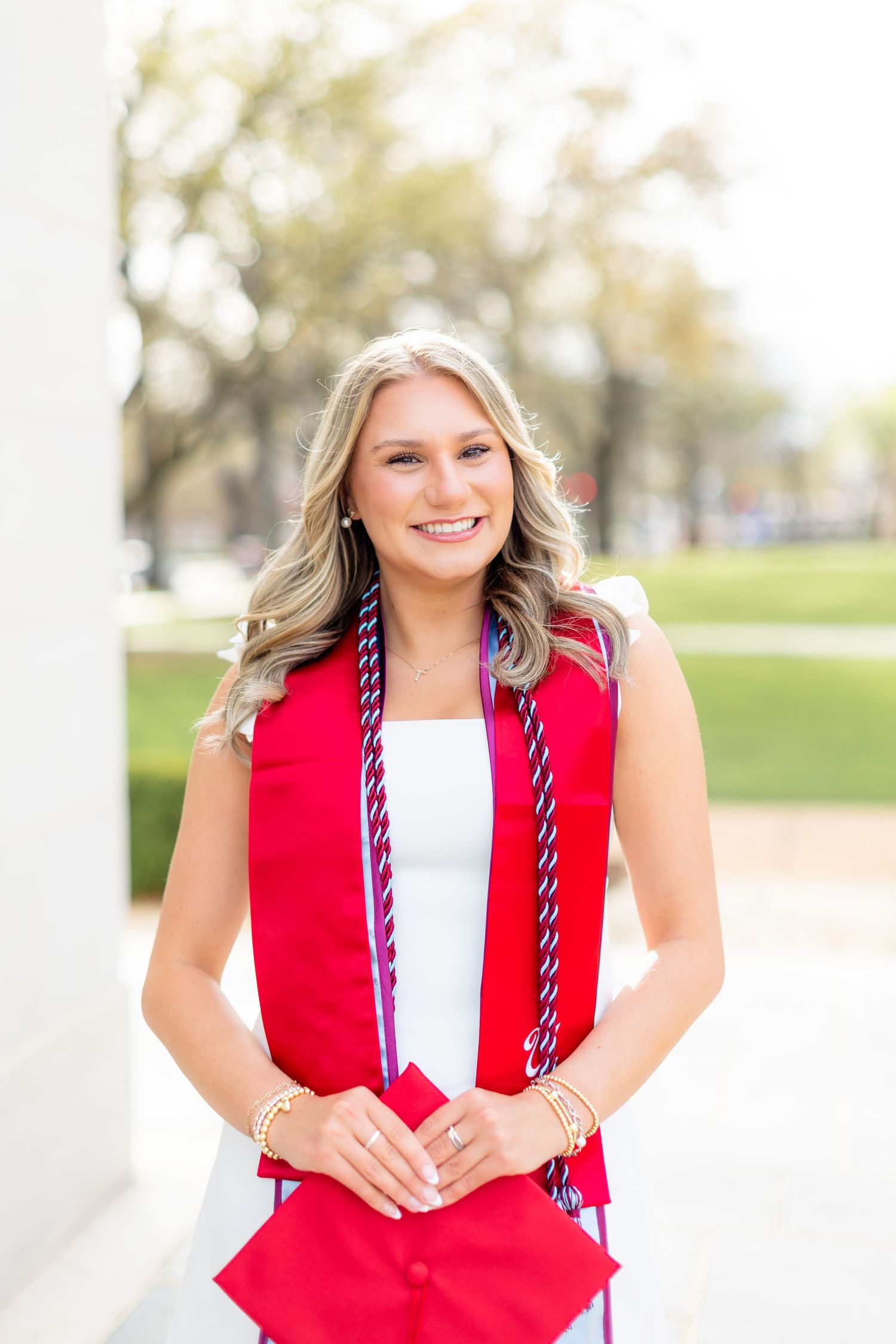 Graduate in red graduation stole and white dress smiles outdoors on sunny day with trees in background.