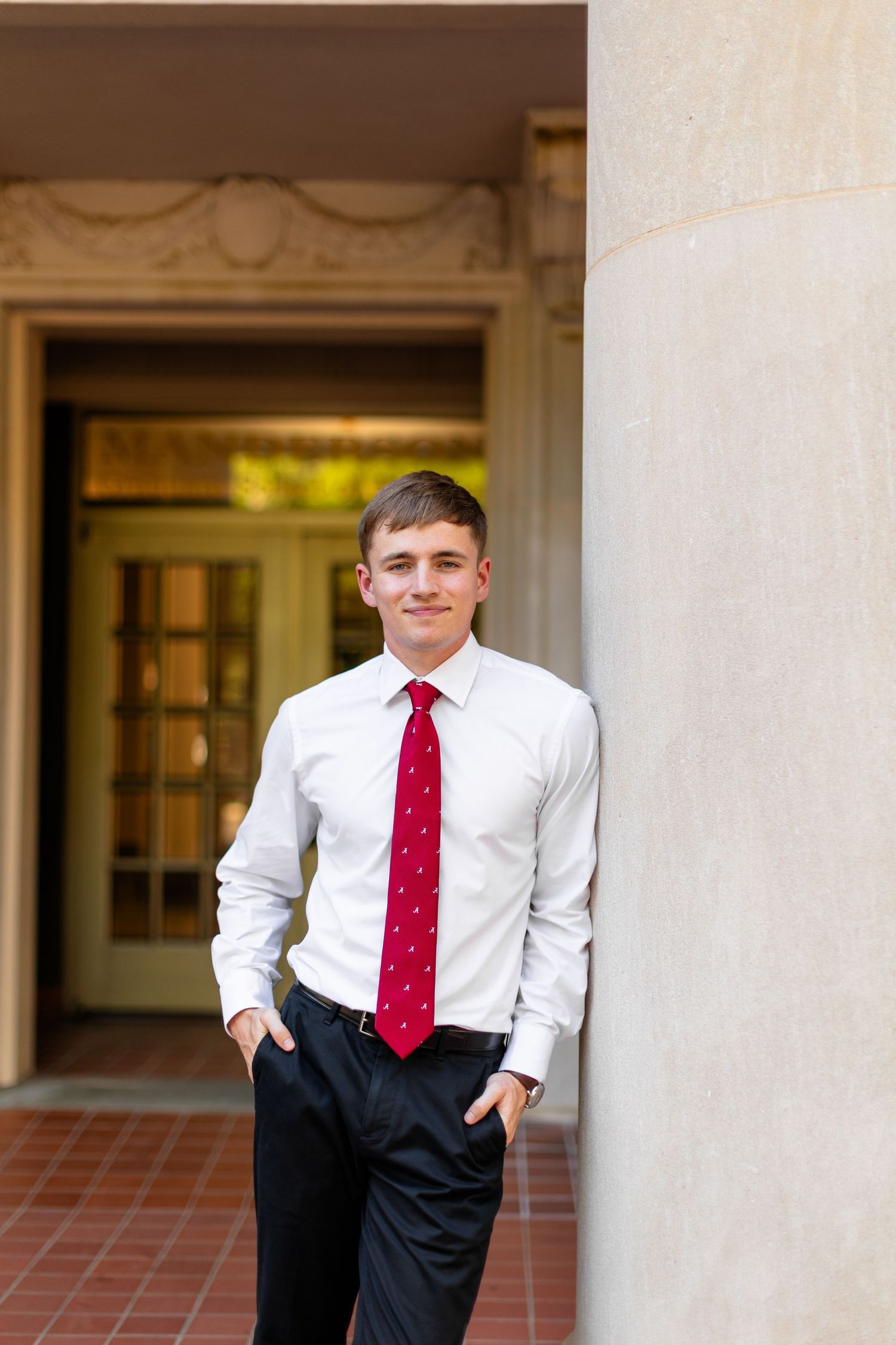 Young professional in white dress shirt and red tie poses casually against a column in an elegant hallway entrance.