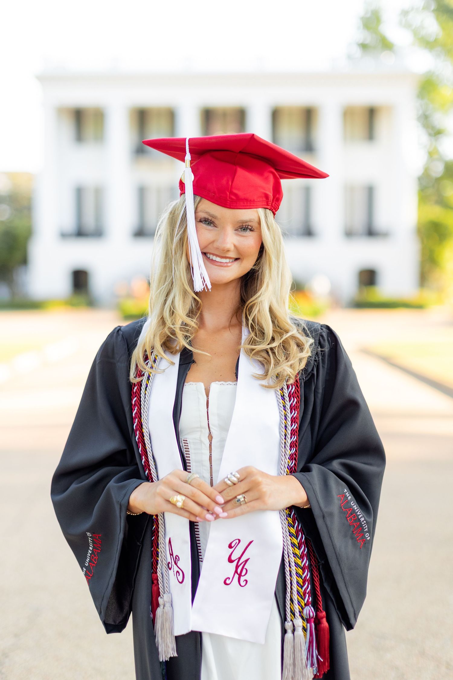 Graduate in academic regalia poses outdoors in front of a white building during commencement.