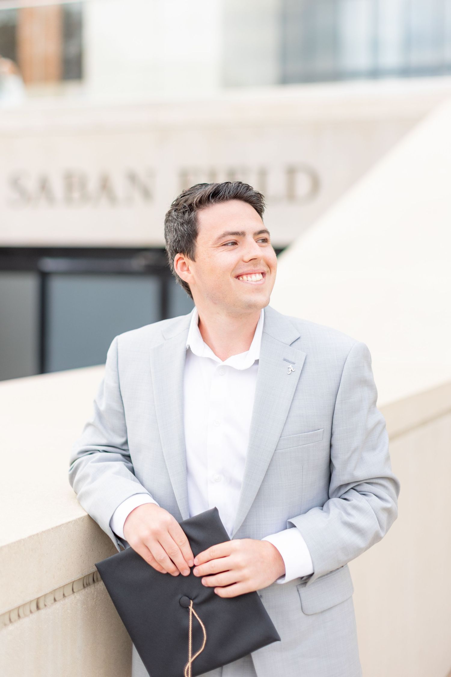 Professional in light gray suit smiles while holding graduation materials outside building.