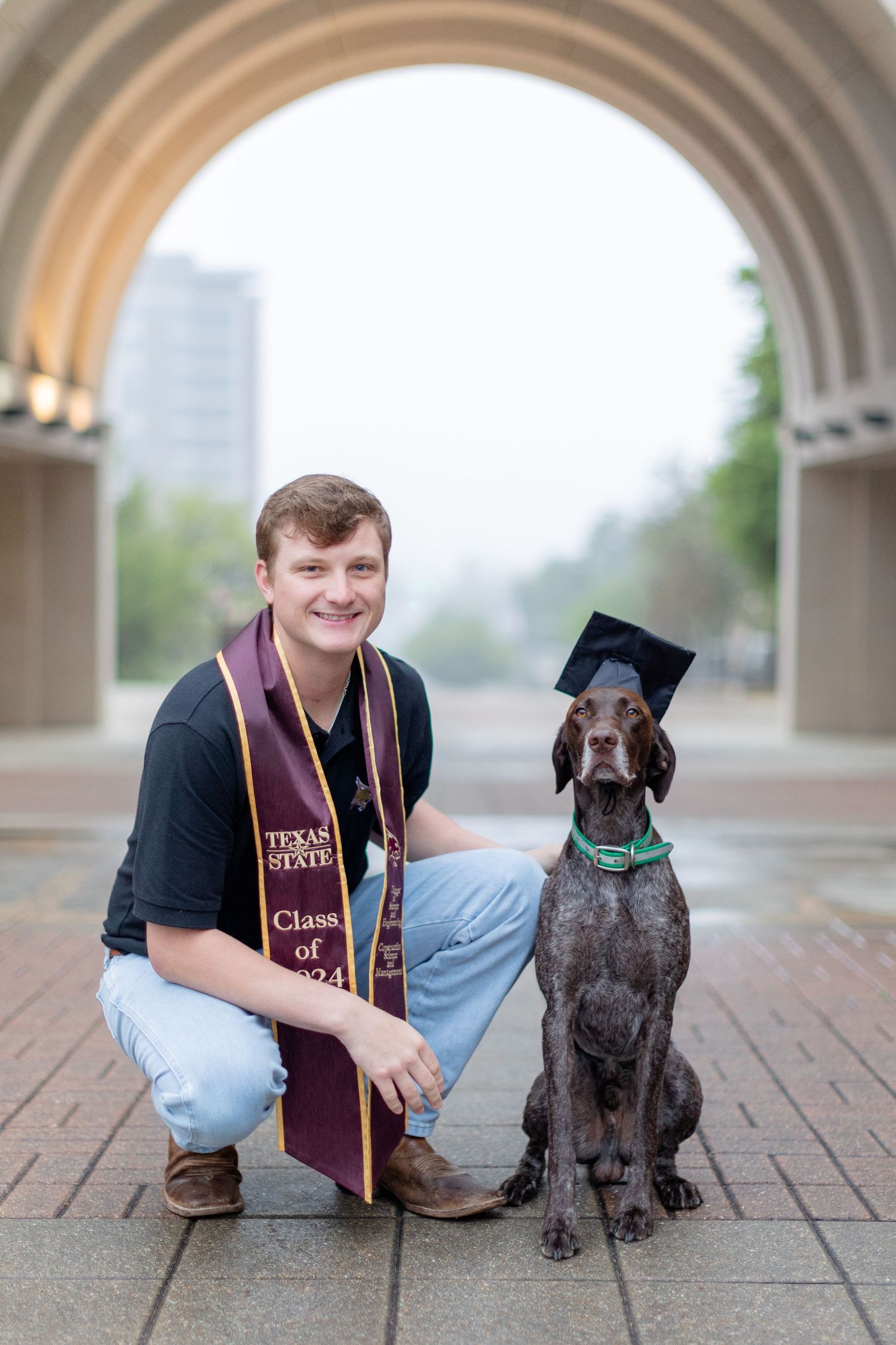 A graduate and their German Shorthaired Pointer dog wearing matching graduation caps under an archway.