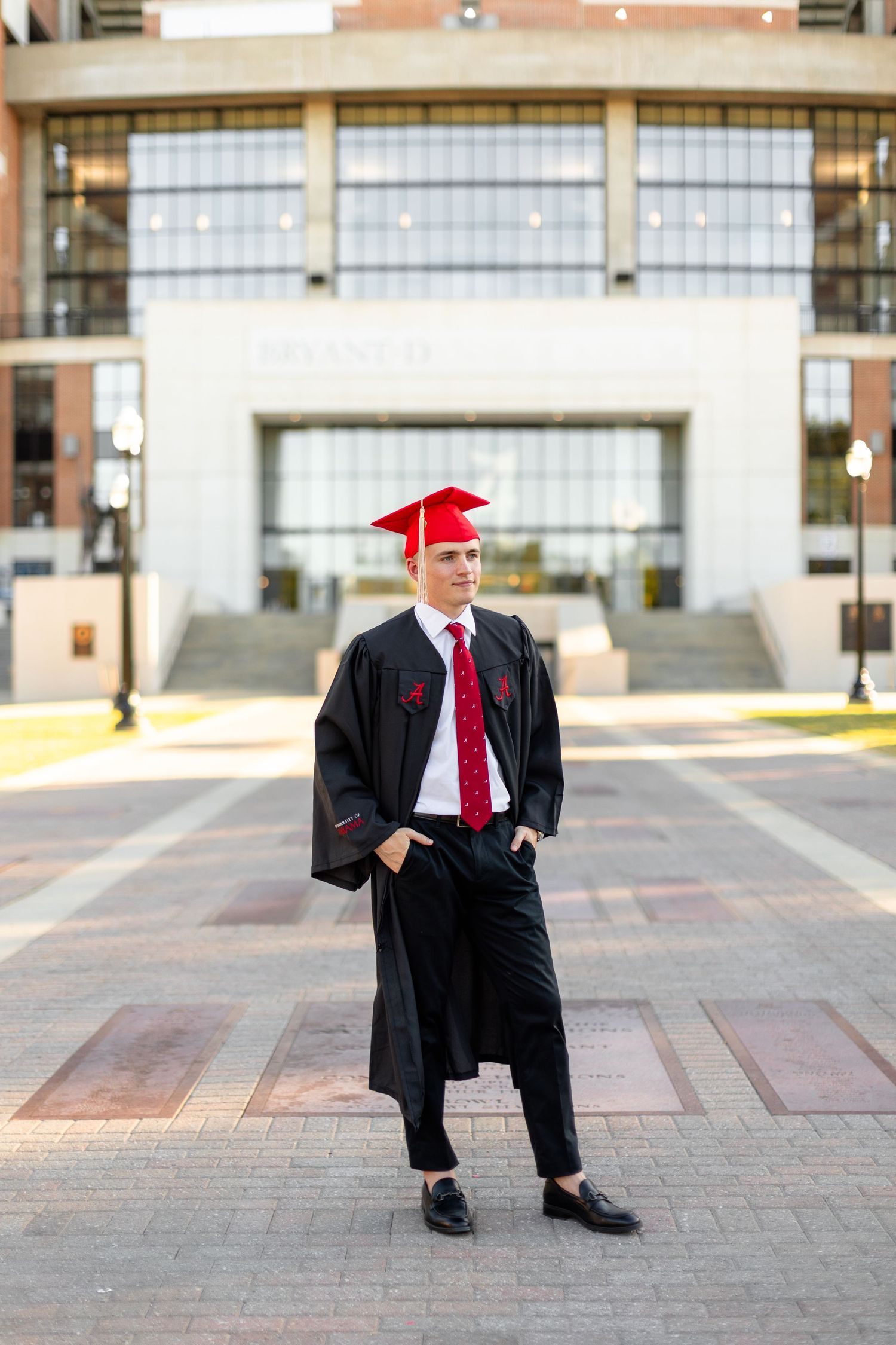 Graduate in cap and gown strikes poses on brick courtyard with modern building in background.