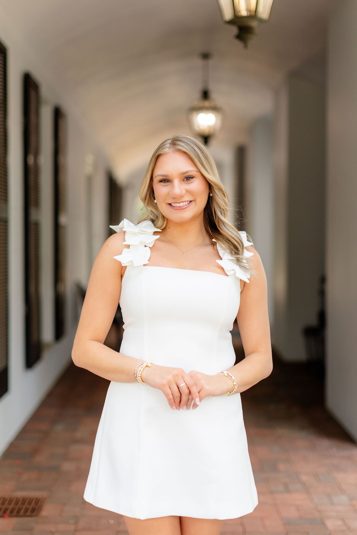 A person wearing a white summer dress with floral shoulder details stands in an elegant covered walkway with pendant lights.