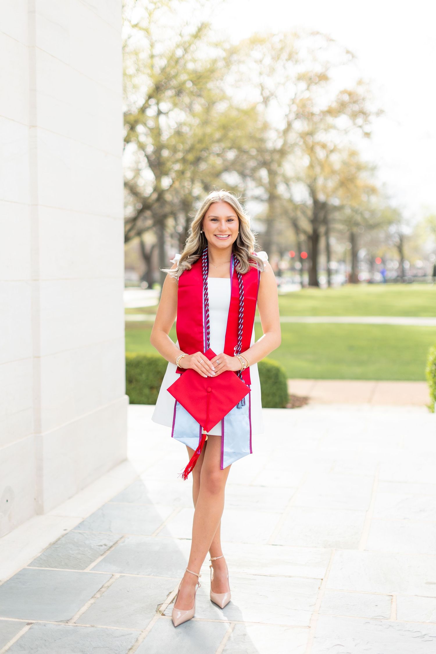 A woman in a red sleeveless blazer and white dress poses outdoors near white columns in a park setting.