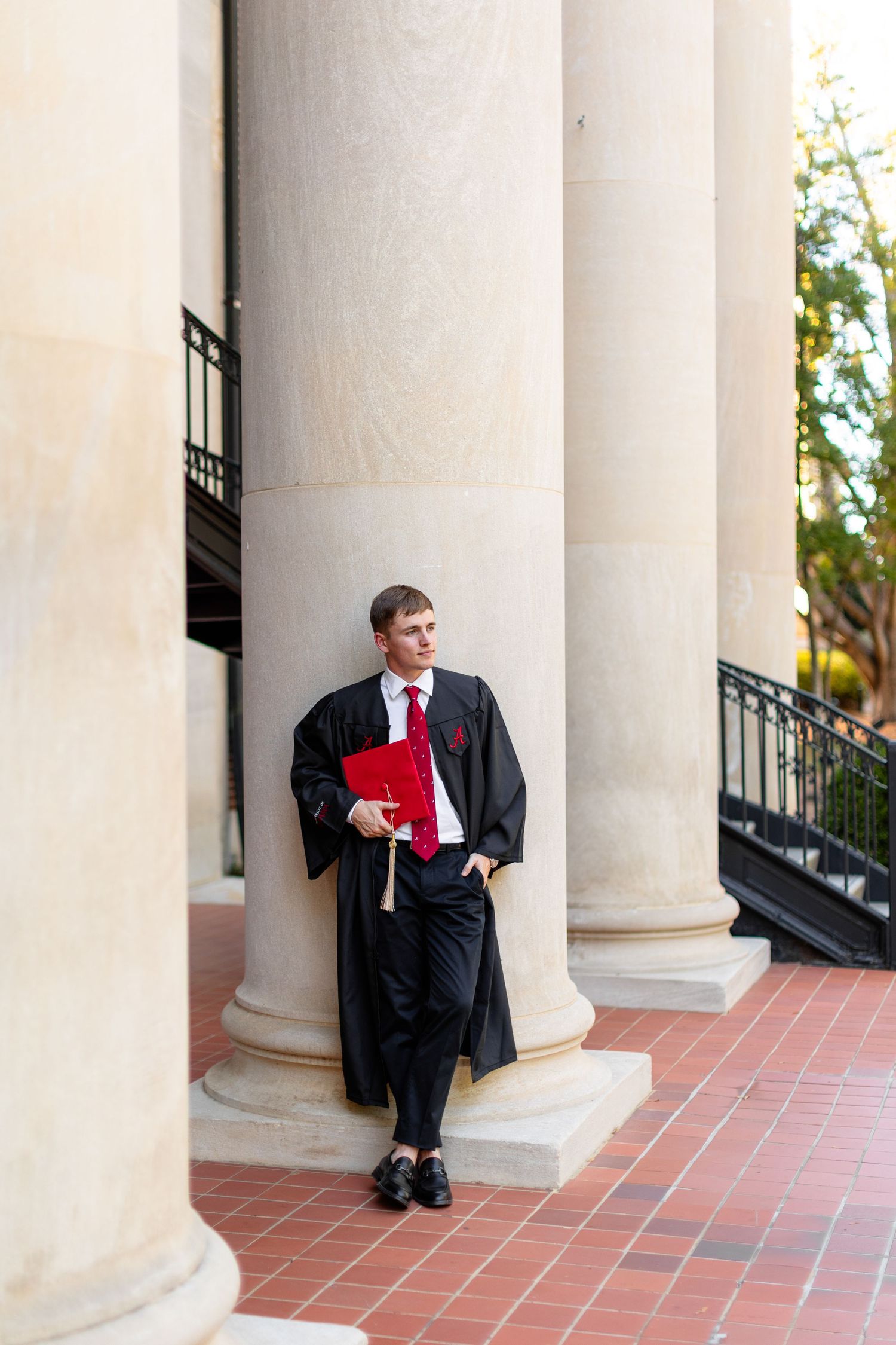 Graduate in academic regalia poses between classical white columns on brick walkway.