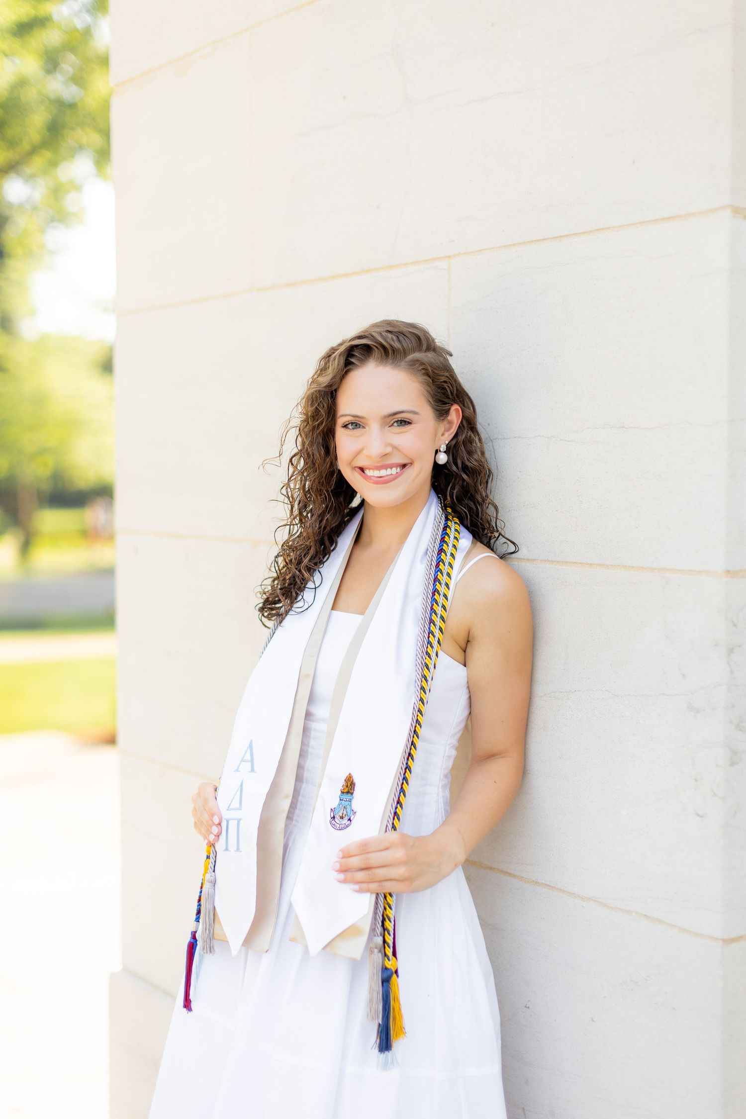 Graduate in white dress with decorative tassels against light wall.