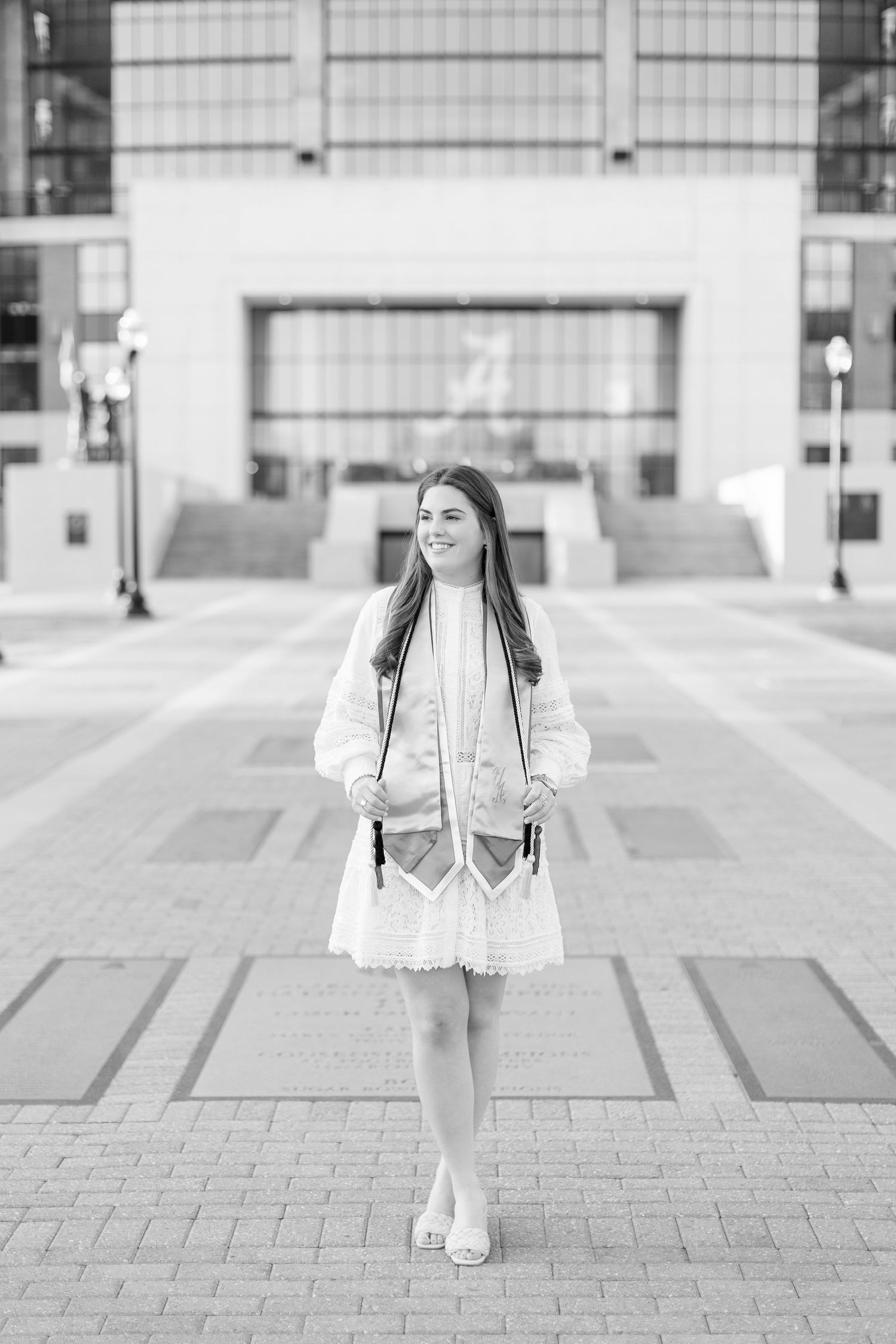 Black and white portrait of someone in a white dress and coat standing on a paved plaza in front of a modern building.