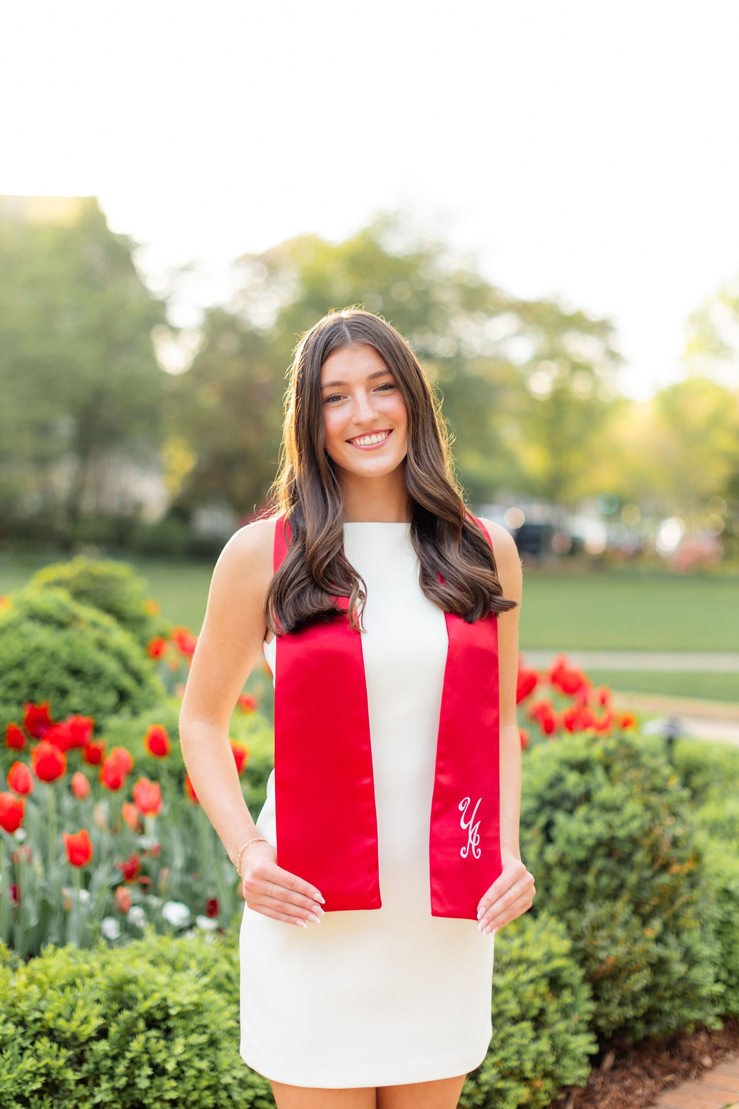 A person in a white top holds a red graduation stole while standing in a garden with blooming red tulips.