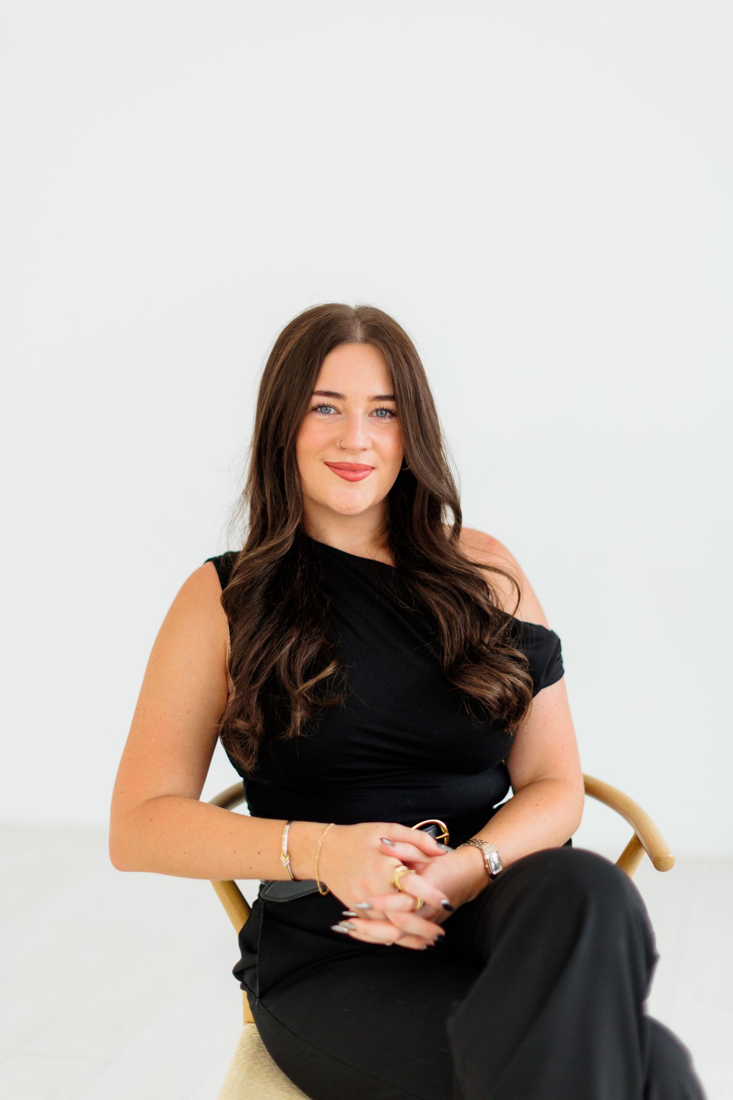 Woman with long brown hair in black asymmetric top sits in wooden chair against white background.
