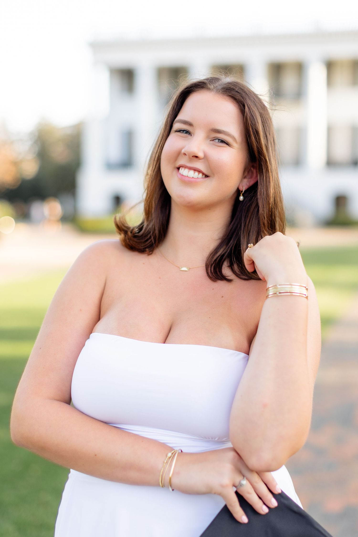 A person in a white strapless top poses outdoors with a bright smile and natural lighting.