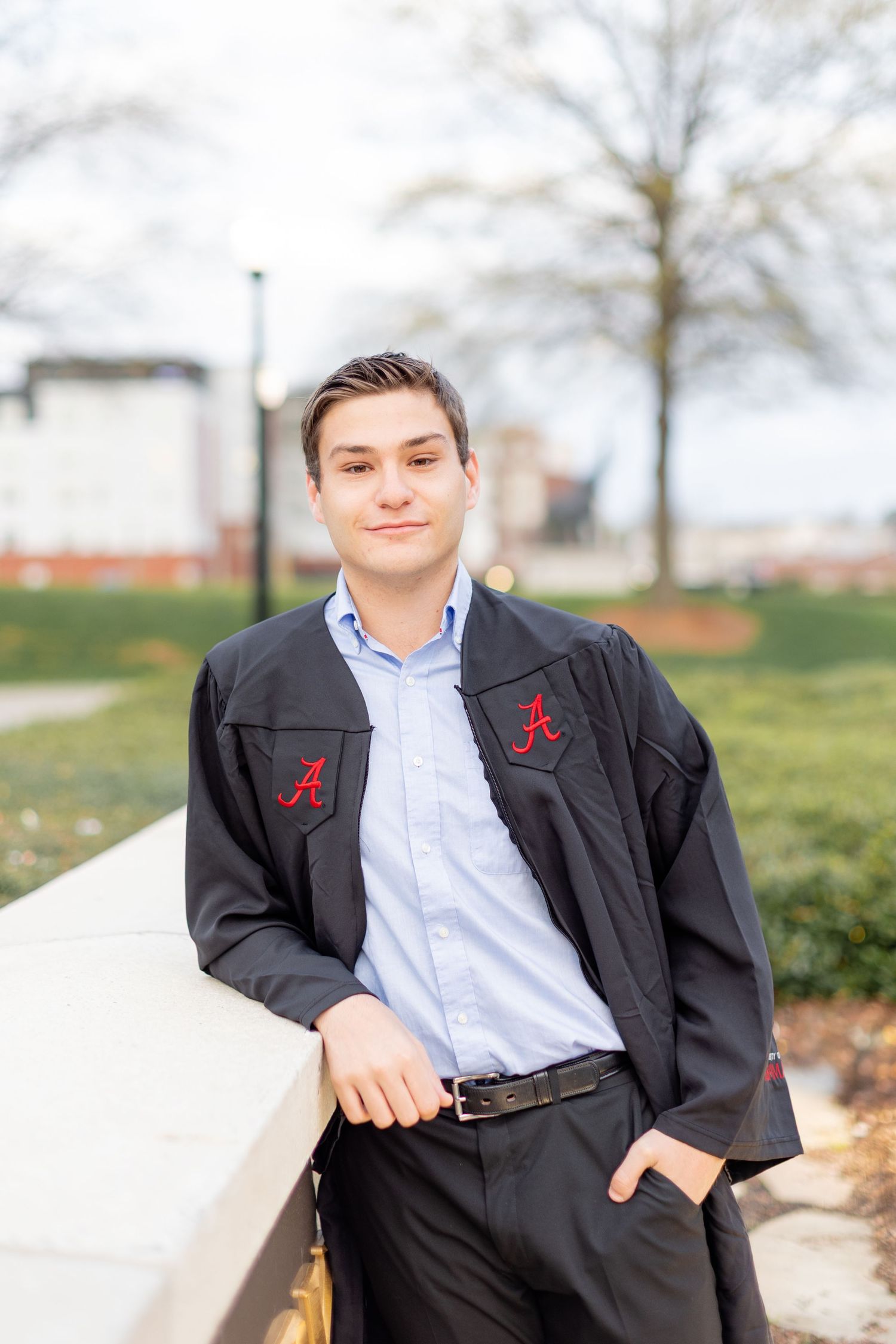 Graduate in academic regalia with Alabama 'A' logo poses outdoors on a sunny day.