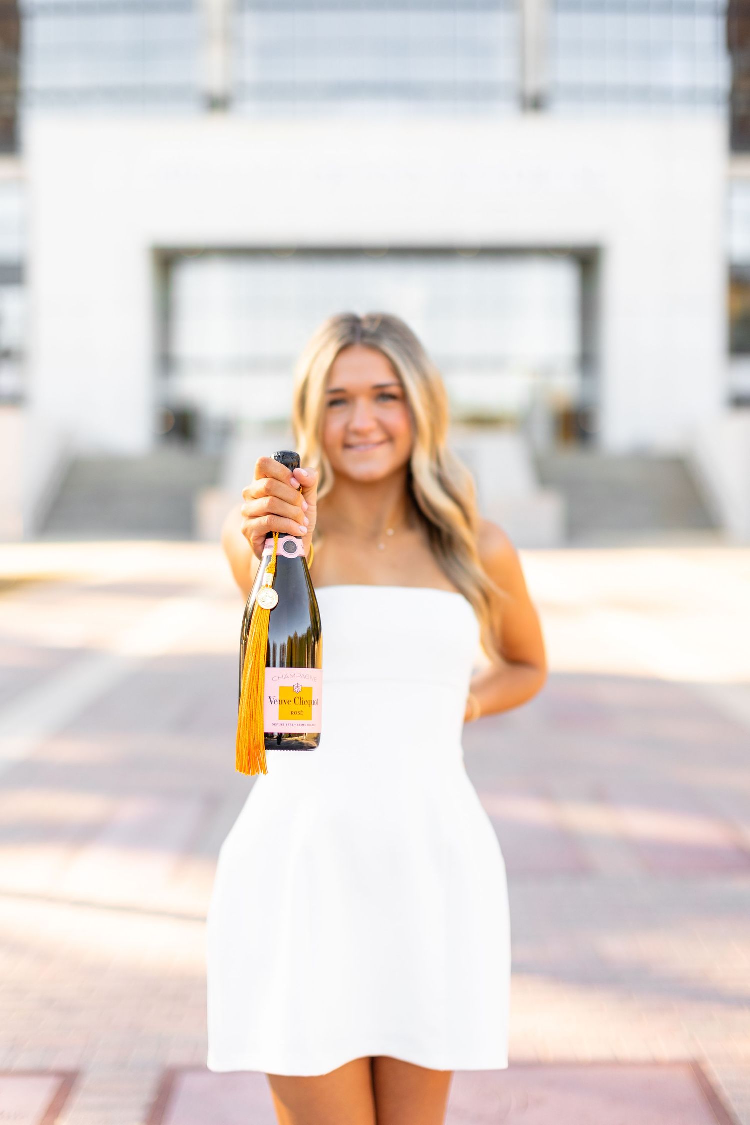 Someone in a white strapless mini dress holds up a bottle of wine while standing outdoors in bright sunlight.