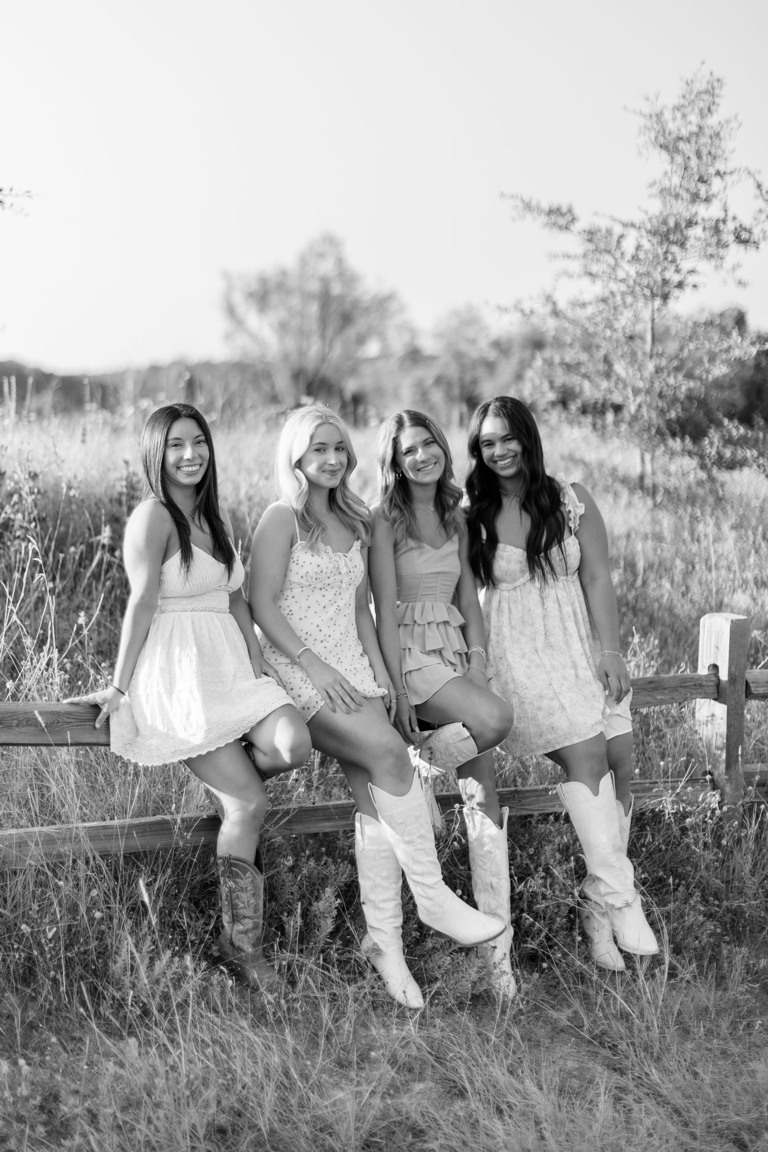 A group of friends in light summer dresses sit together on a rustic wooden fence in a black and white photograph.