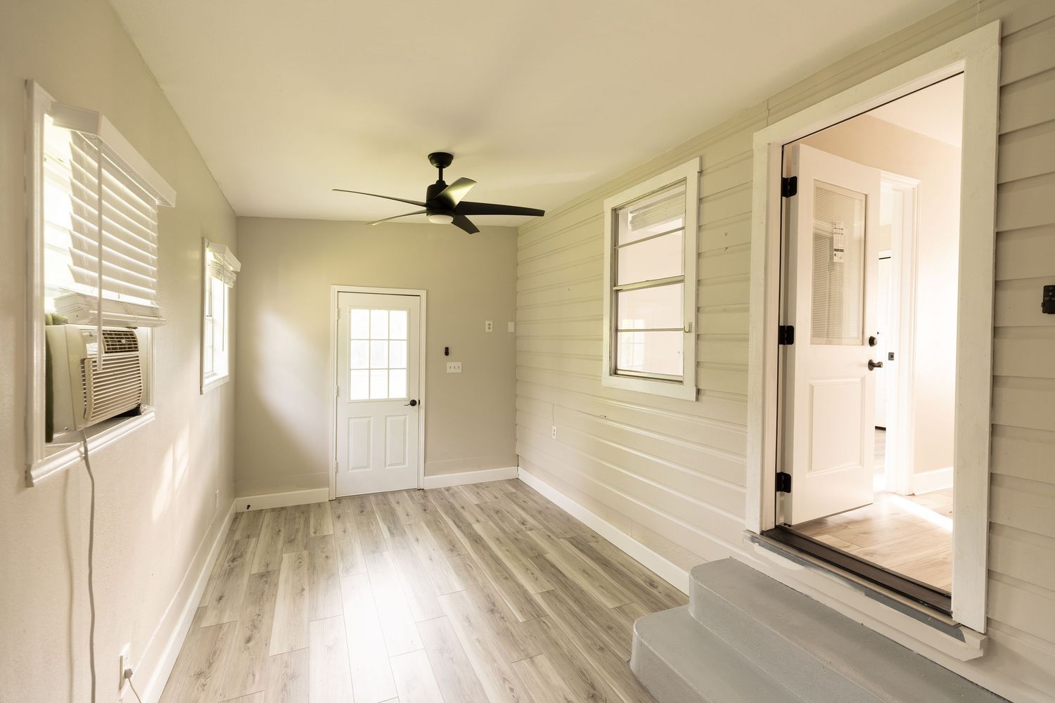Bright sunroom with shiplap walls, ceiling fan, hardwood floors, window AC unit, and door leading to adjacent room.