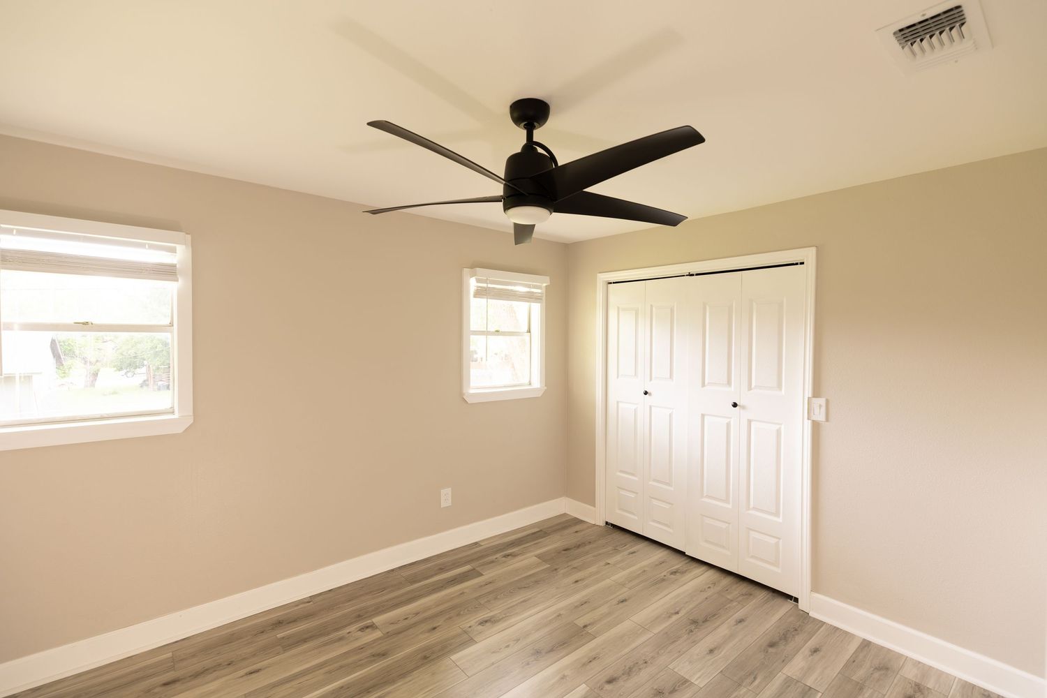 Empty bedroom with gray hardwood floors, white closet doors, two windows, and a black ceiling fan.