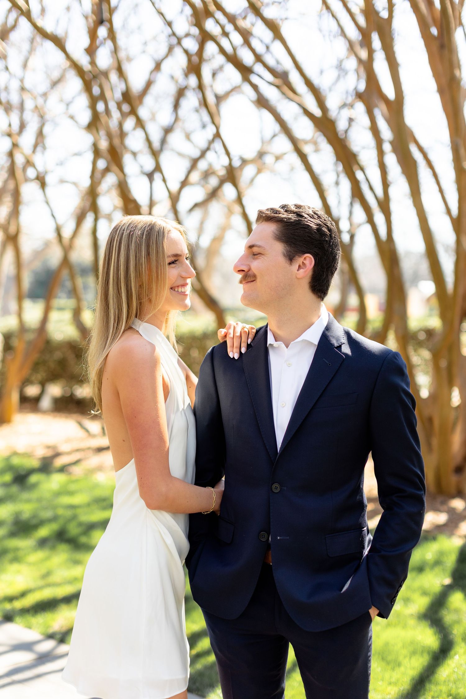 A couple shares joyful laughter together during a sunny outdoor photoshoot among trees.