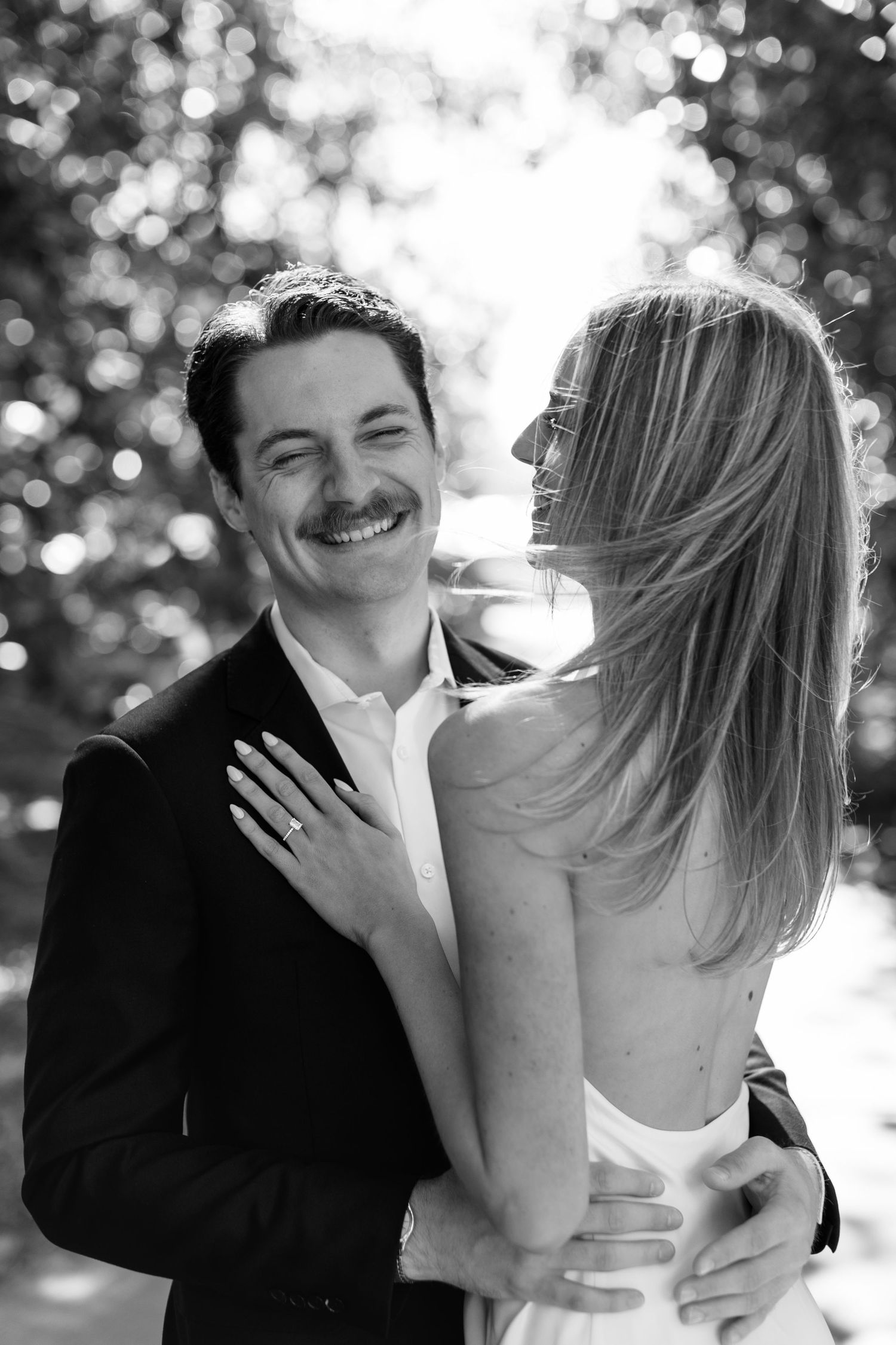 A black and white romantic outdoor portrait of a couple embracing and laughing together in natural lighting.
