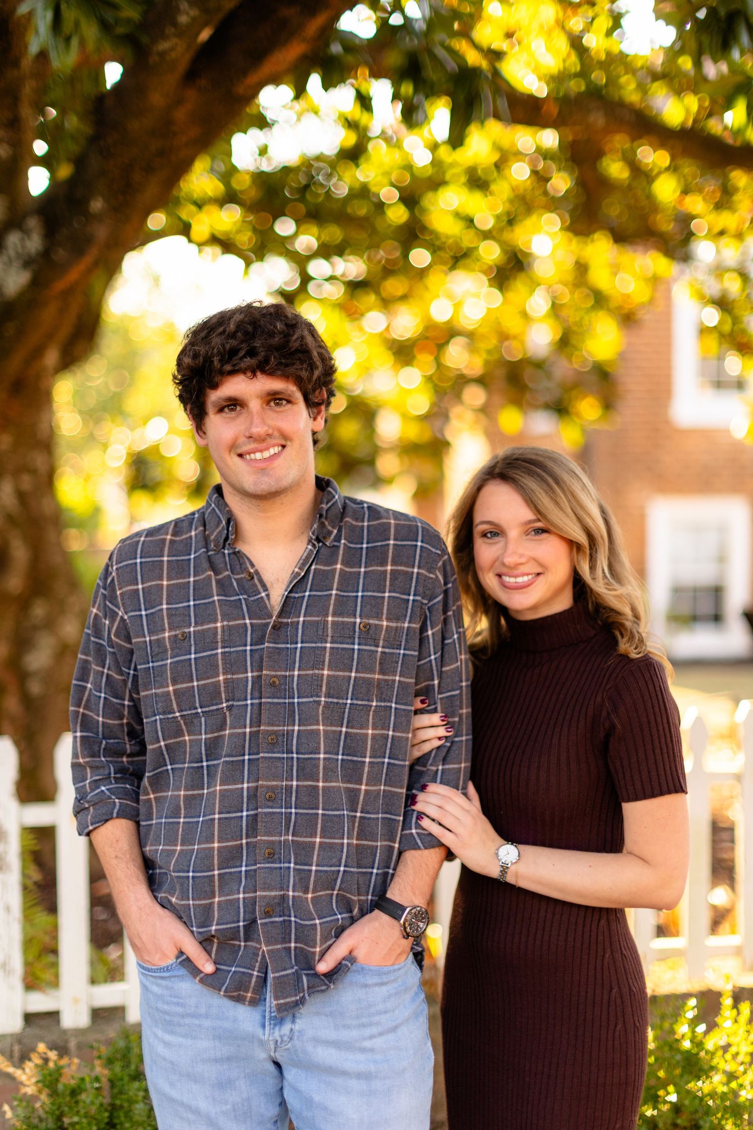 A couple stands together outdoors in autumn, with a brown sweater dress and plaid shirt against golden bokeh lighting.