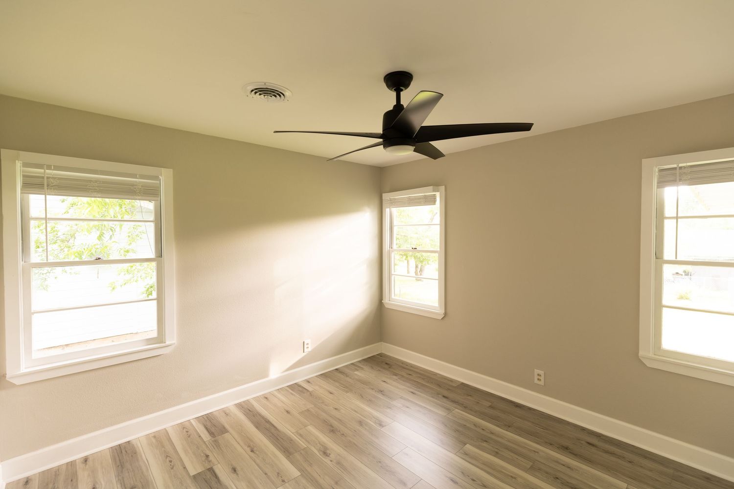 Bright empty room with hardwood floors, two windows, and a black ceiling fan.